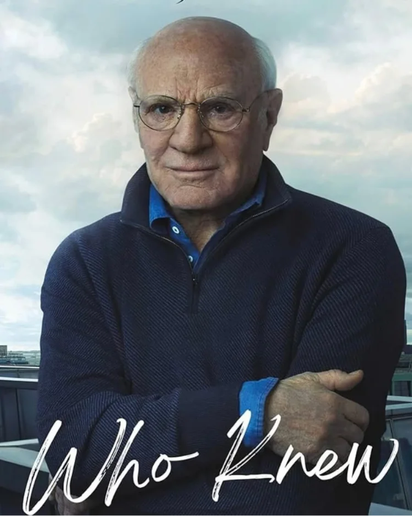 Barry Diller looks at the viewer, wears glasses, and crosses his arm over his body, holding his left arm, with blue sky and white clouds, and a city behind him, and the name of his memoir, Who Knew in white handwritten cursive letters