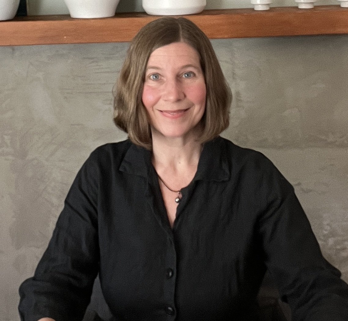 A woman with shoulder-length brown hair, wearing a black shirt, sitting indoors with a neutral-colored wall behind her and a wooden shelf with white decorative objects above.