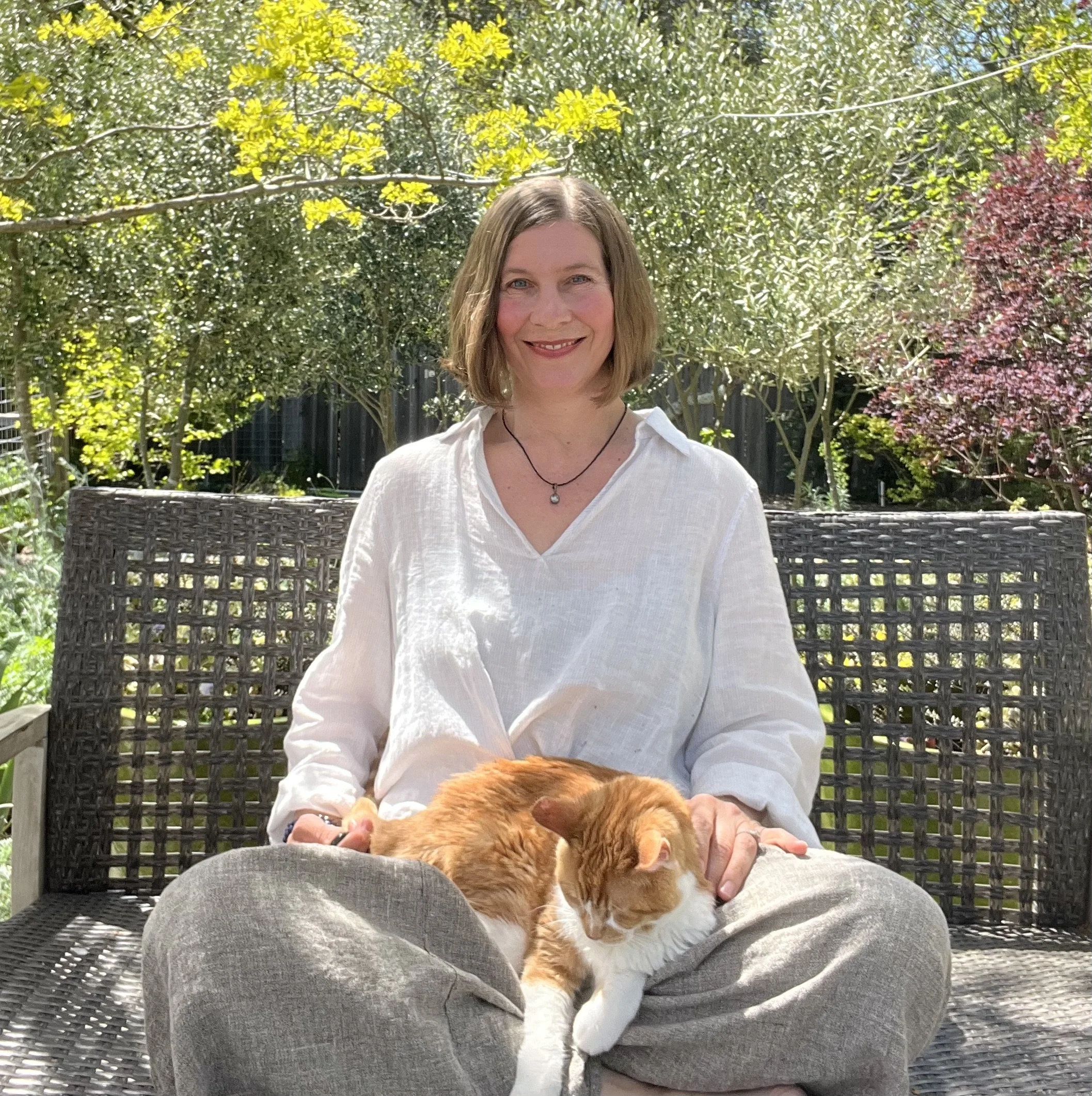 Woman sitting cross-legged on outdoor bench with orange and white cat resting on her lap, surrounded by greenery and trees in a backyard on a sunny day.