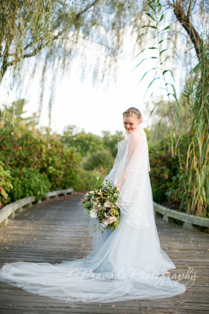 A bride standing on a wooden bridge holding a bouquet in a lush, green outdoor setting with trees and bushes.