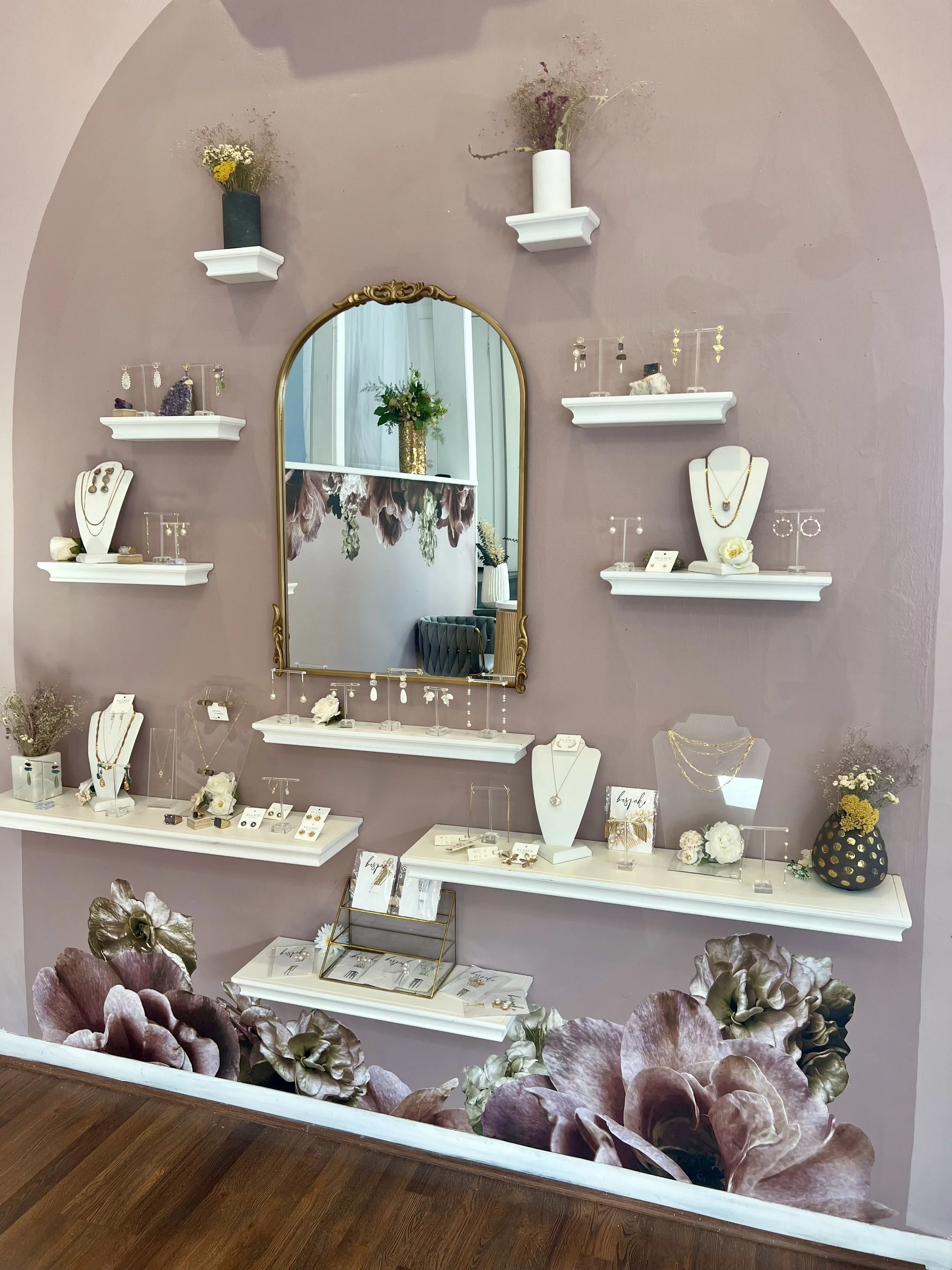 Jewelry display wall with shelves holding earrings, necklaces, and bracelets, a mirror, and floral decorations on a pink wall.