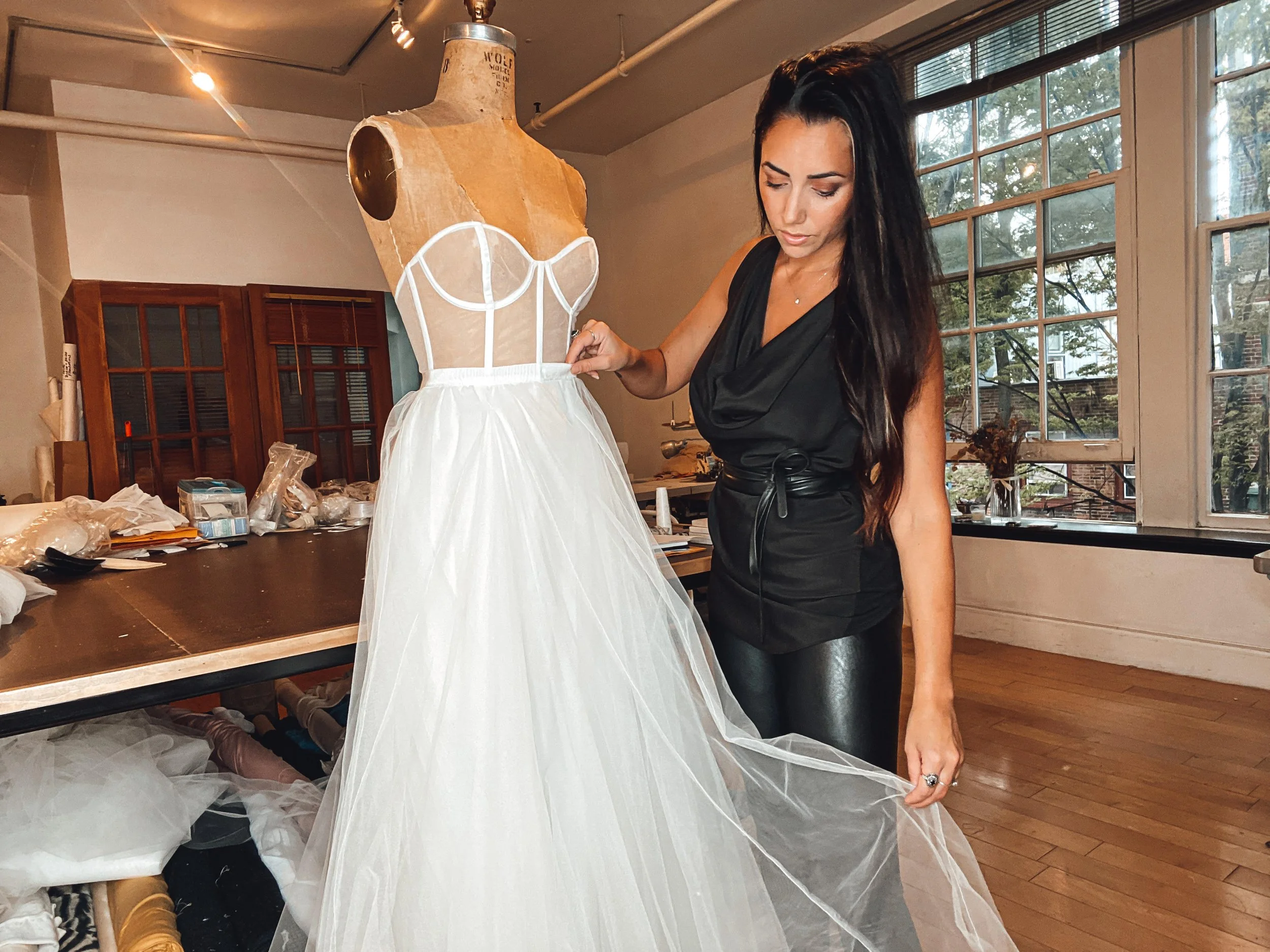 Woman with long dark hair adjusting a white tulle wedding dress on a mannequin in a well-lit room with large windows.