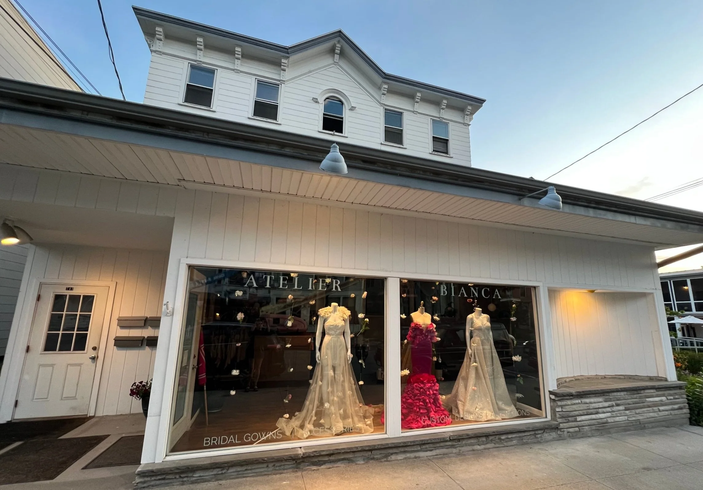 Storefront window display with bridal gowns and a pink dress, sign reading 'Atelier Bianca', on a street with a white building and sidewalk.