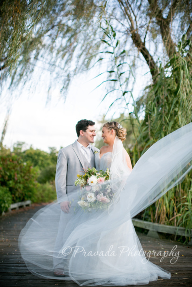A bride and groom standing on a wooden path outdoors, smiling at each other, with trees and greenery in the background. The bride holds a large bouquet, and her veil is flowing in the wind.