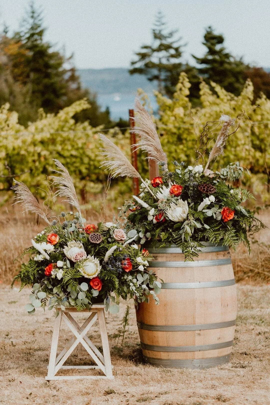 Two large floral arrangements with various flowers and greenery, one inside a wooden barrel and the other on a small stand, set outdoors in a vineyard with trees in the background.