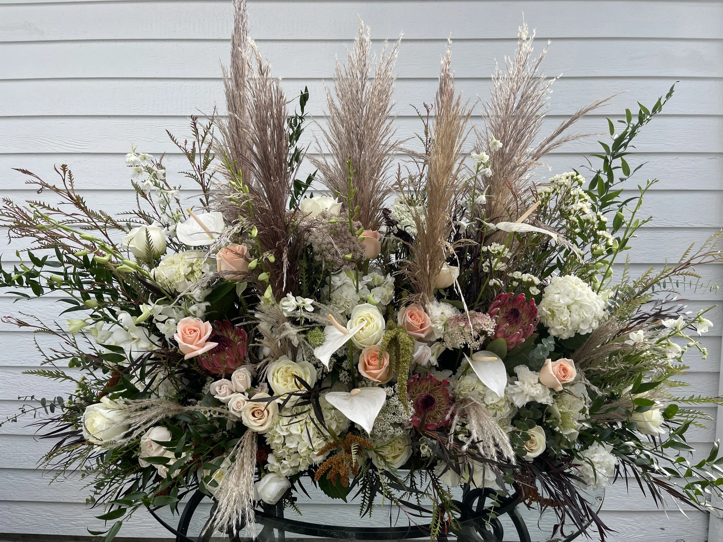 A large floral arrangement featuring white, cream, and blush roses, white hydrangeas, white lilies, purple proteas, and various greenery, with tall, wispy pampas grass accents, set against a white, horizontal-slat background.