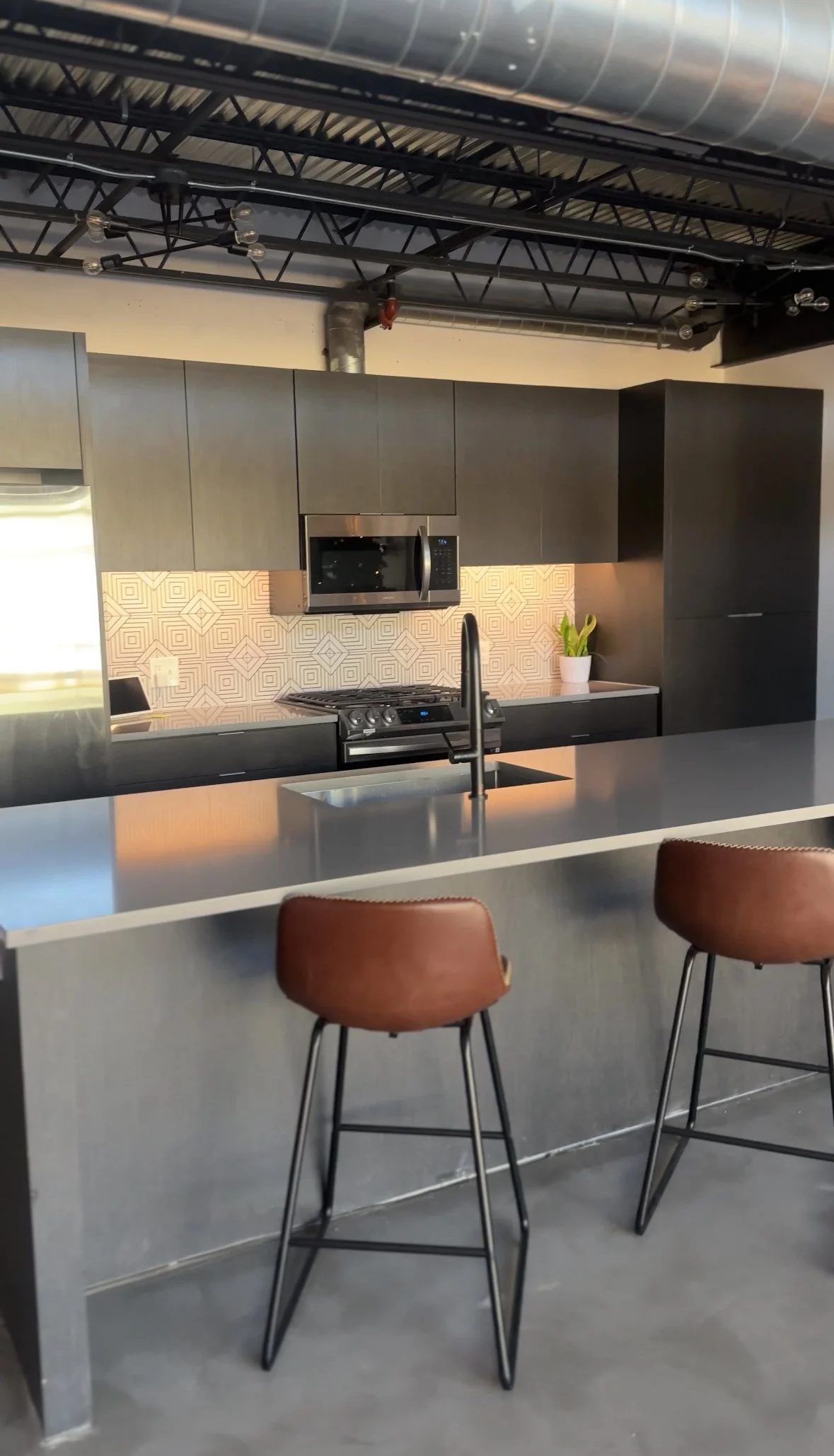 Modern kitchen with dark cabinetry, stainless steel appliances, a tiled backsplash, a kitchen island with a sink, brown bar stools, and an industrial ceiling with exposed ducts and beams at a photography studio in San Antonio, Texas.