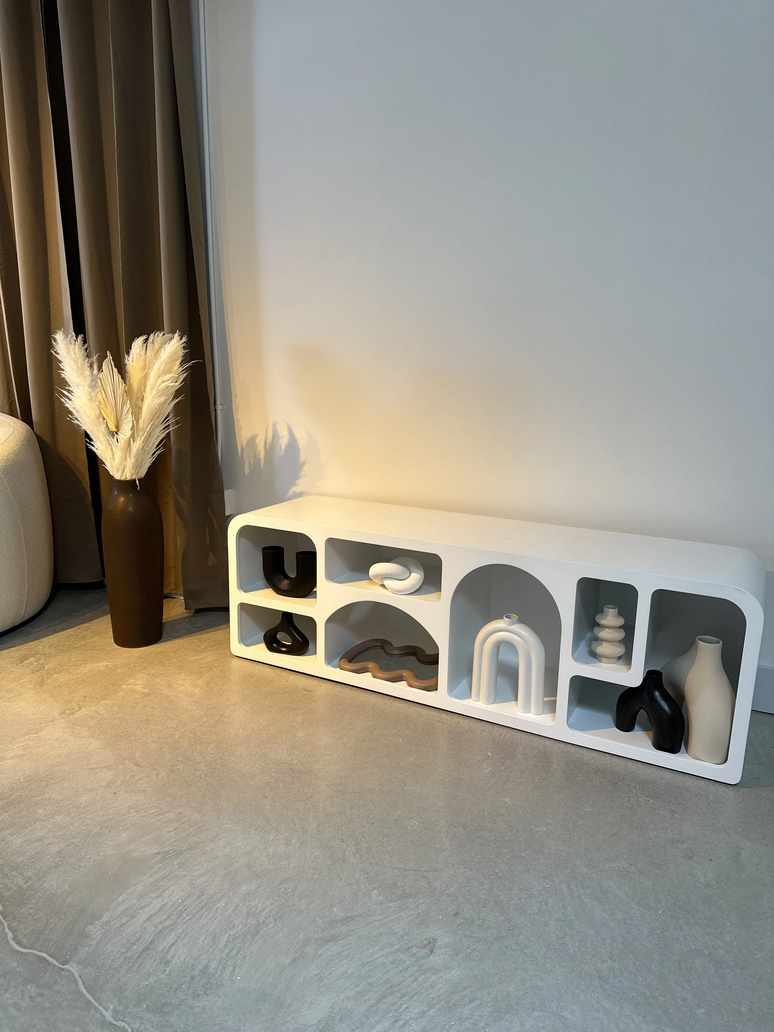 Modern white decorative shelf with abstract black and beige vases and sculptures, positioned on beige carpeted floor next to a dark vase holding pampas grass, near a beige sofa and a window with beige curtains.