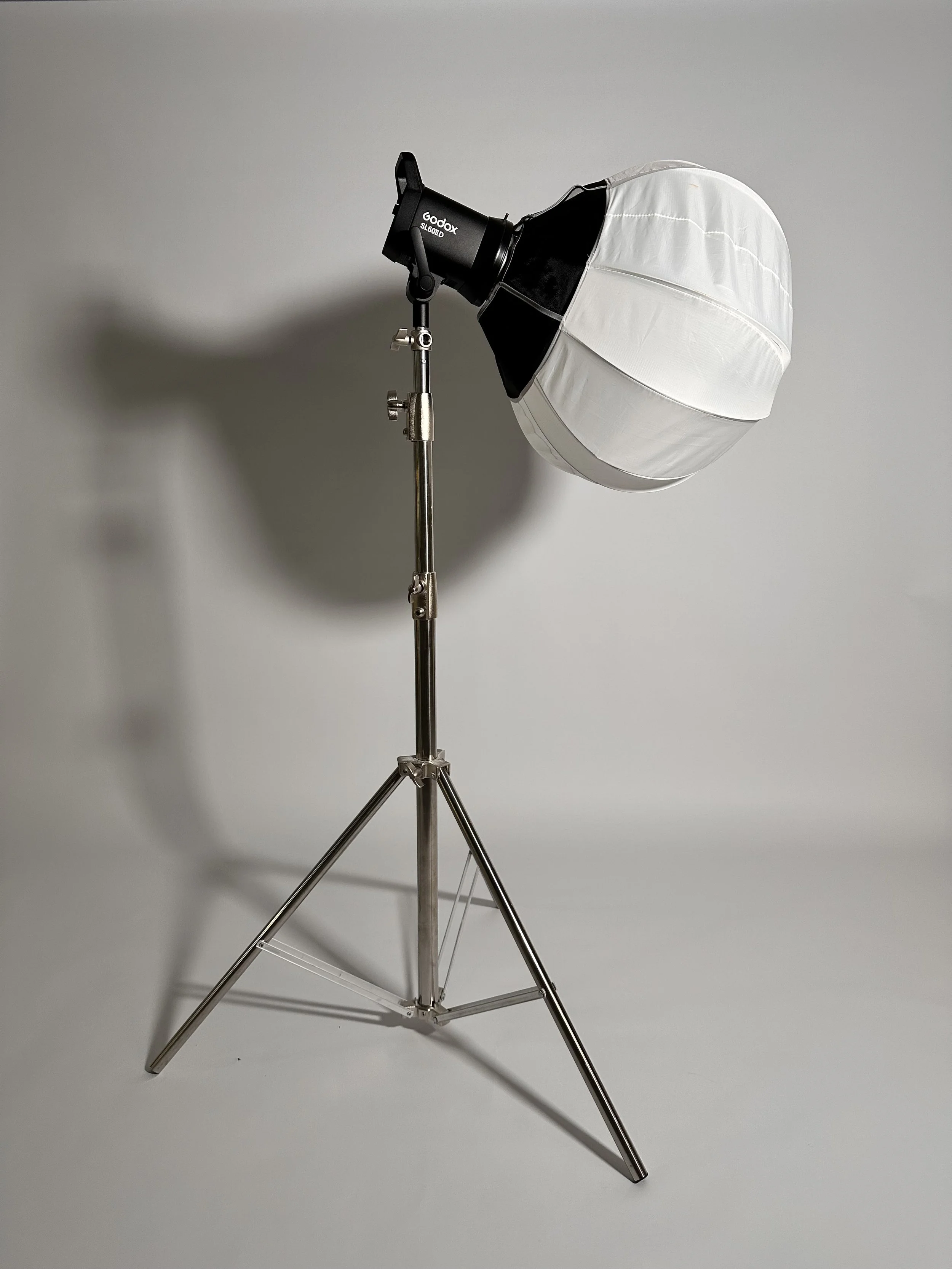 A studio light with a large white diffuser on a metal stand against a plain gray background.