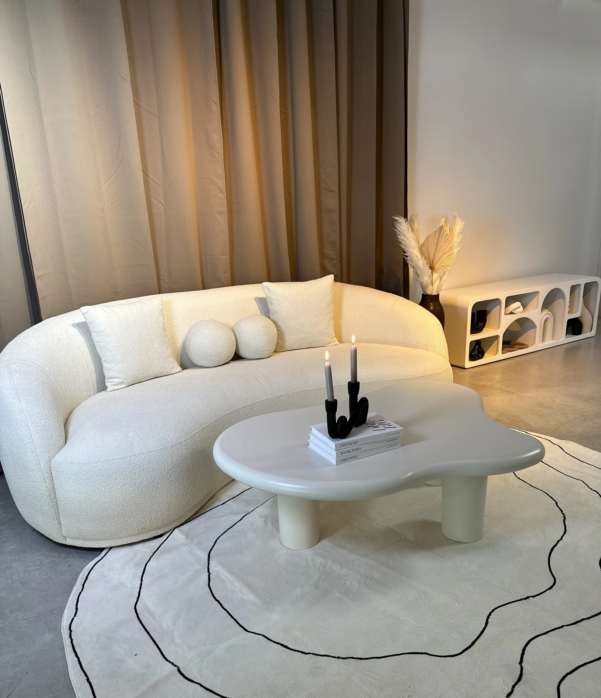 Modern living room with a curved white sofa, decorative pillows, black candle holder with lit candles, a white oval coffee table on a white rug with black lines, a beige cabinet with decorative objects, and a dark vase with pampas grass near beige cu
