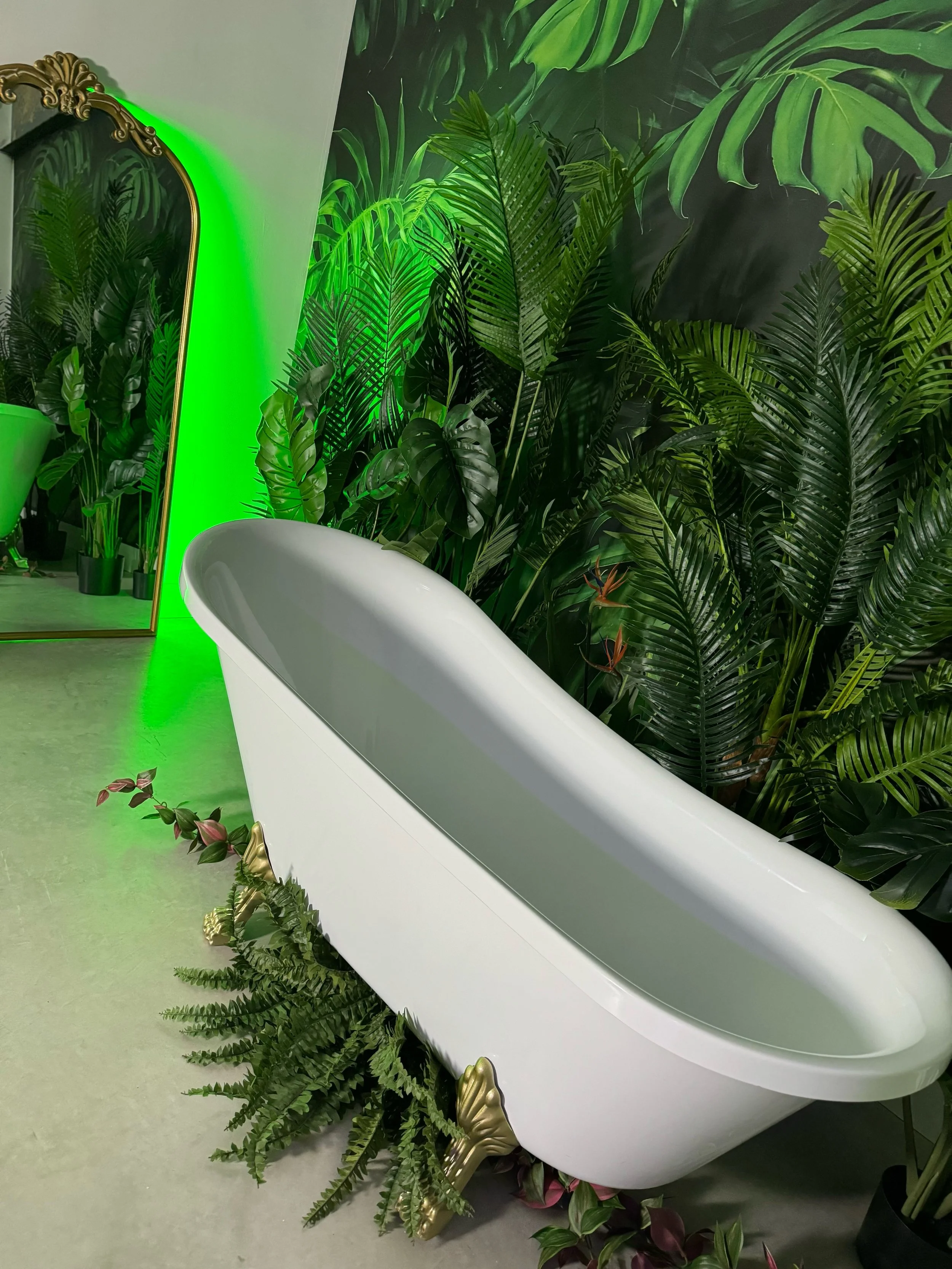 White bathtub surrounded by lush green plants with a mirror reflecting some of the plants and a green light in the background.