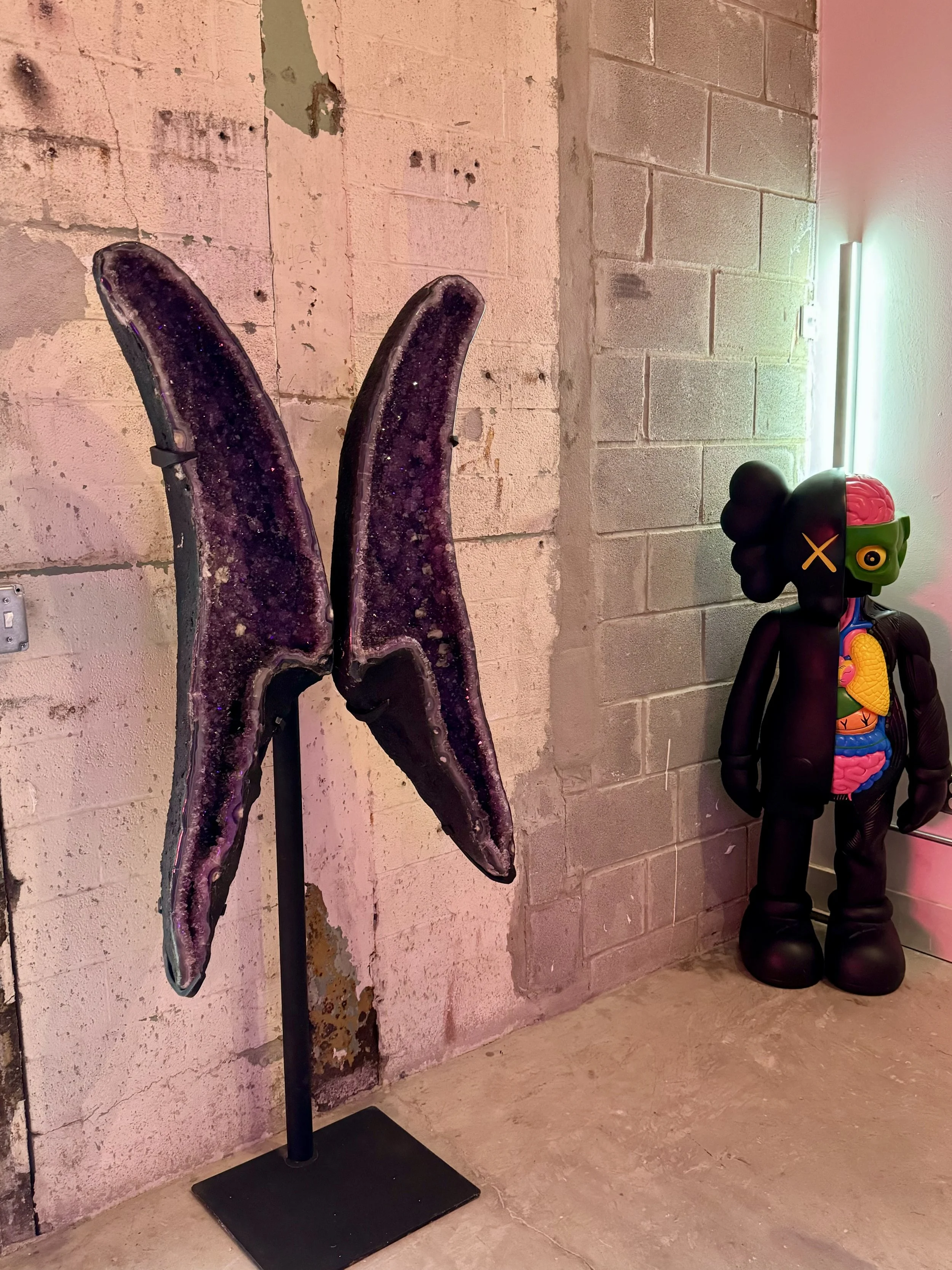 A pair of purple, glittery banana-shaped sculptures on a black stand next to a colorful, artistic robot figure with a visible brain and various patterns on its body.