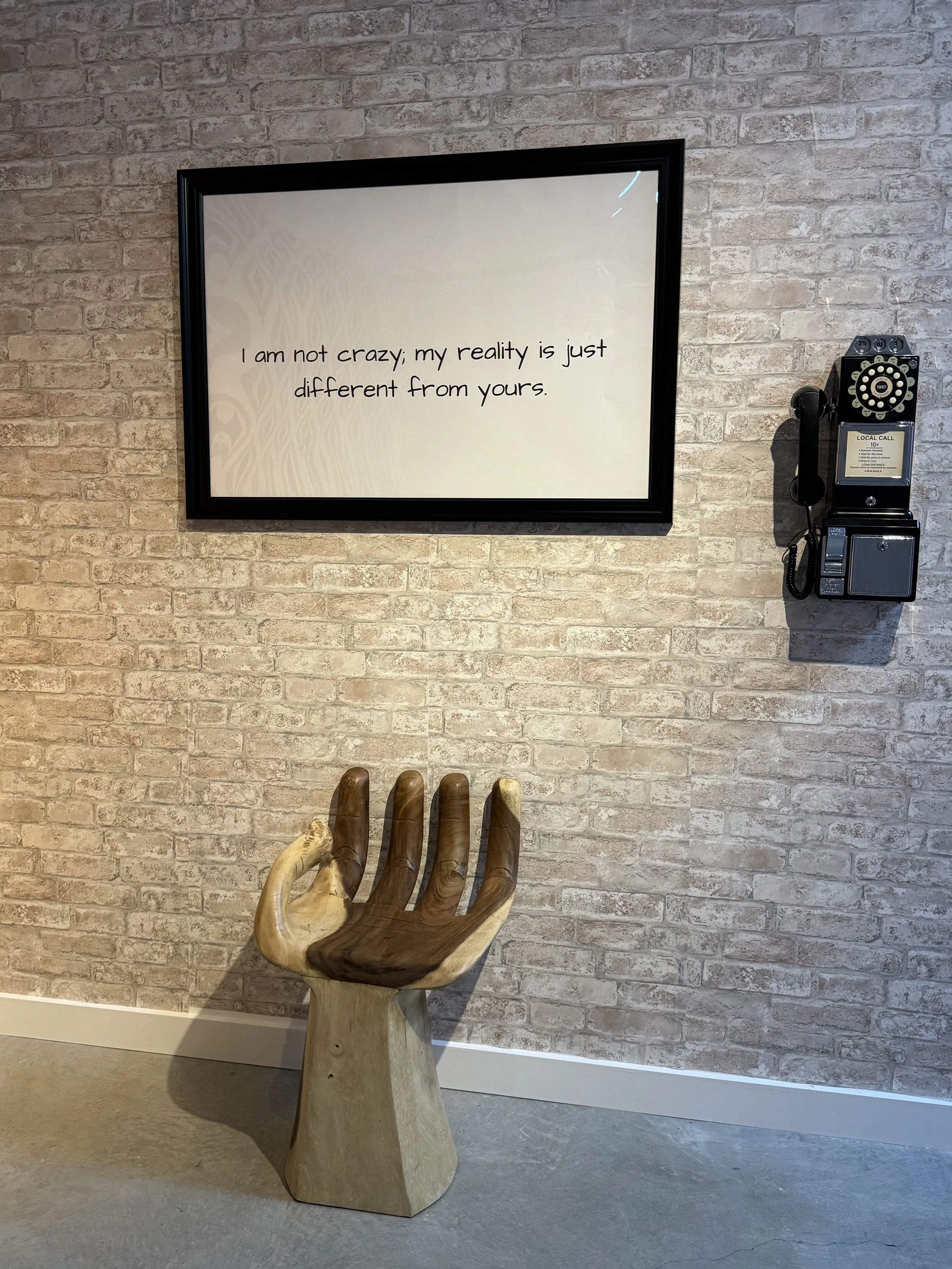 Decorative wall with a framed quote reading, "I am not crazy; my reality is just different from yours," a vintage wall phone, and a wooden hand-shaped chair.