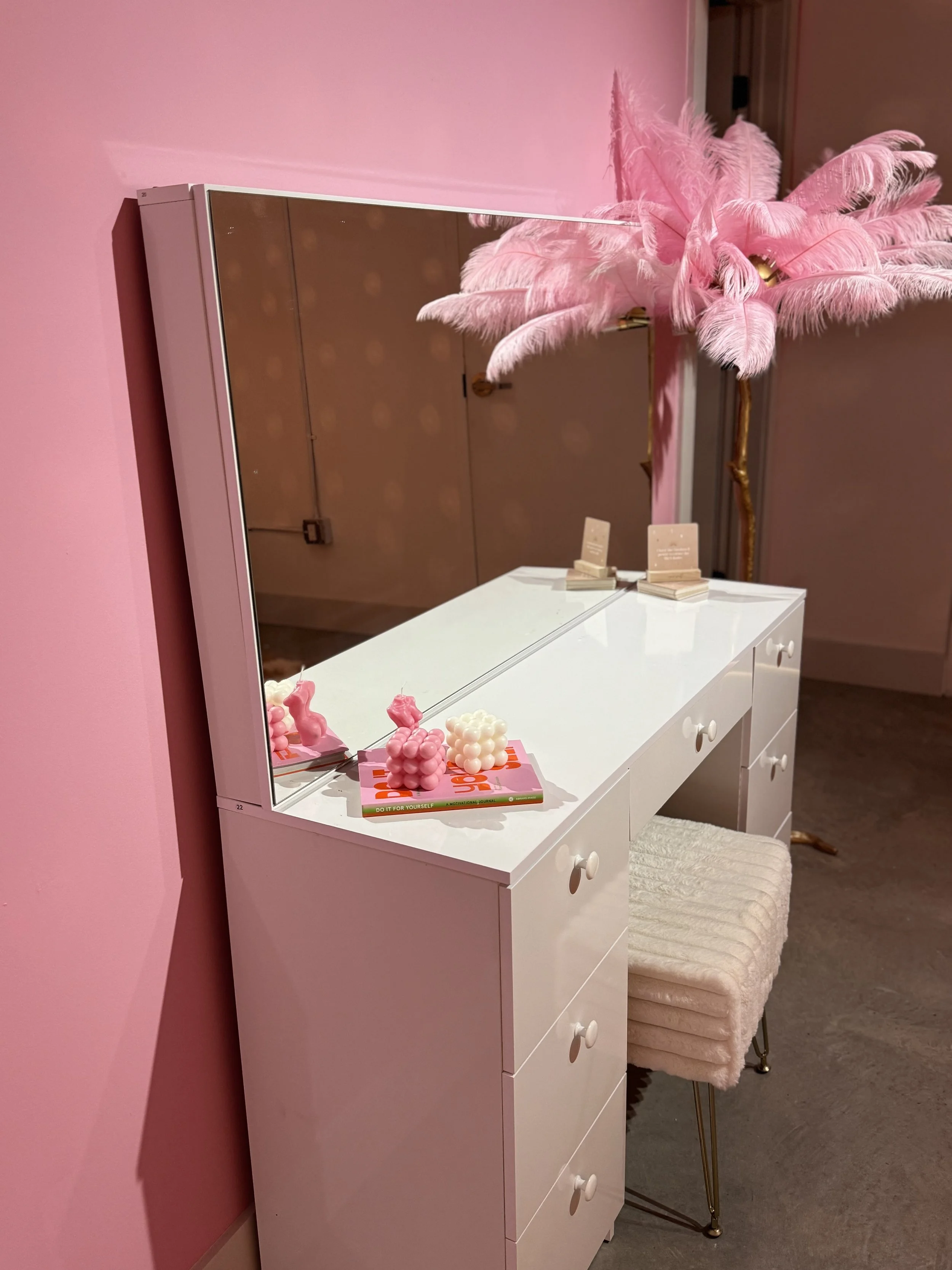 A white vanity table with a large mirror, decorated with pink bubble candles and pink feathered decor on top. The table is against a pink wall and has a plush, cream-colored stool with gold legs in front of it.