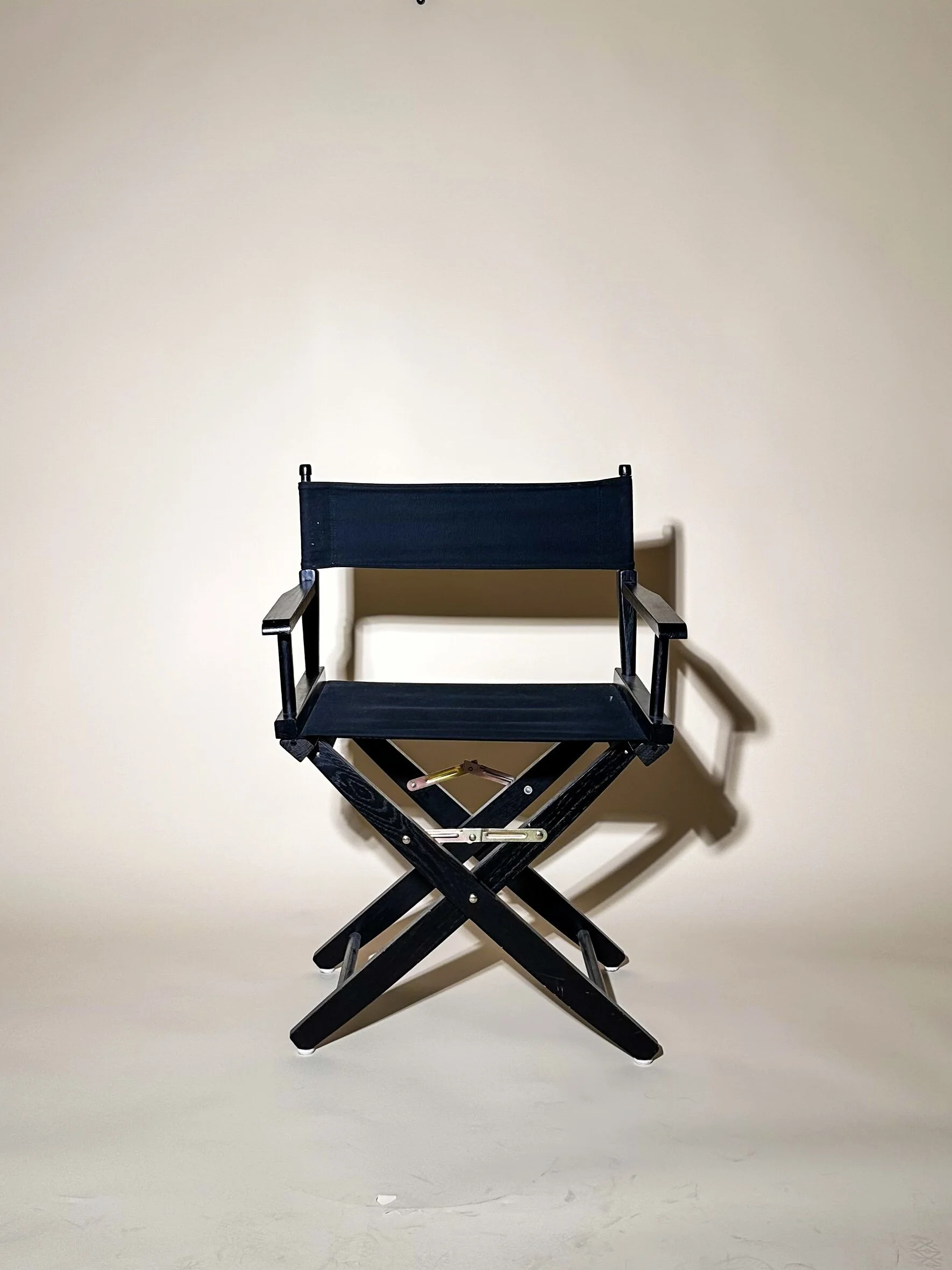 A portable black director's chair with a fabric seat and backrest, set against a plain beige background, casting a shadow on the wall at a photography studio in San Antonio, Texas.