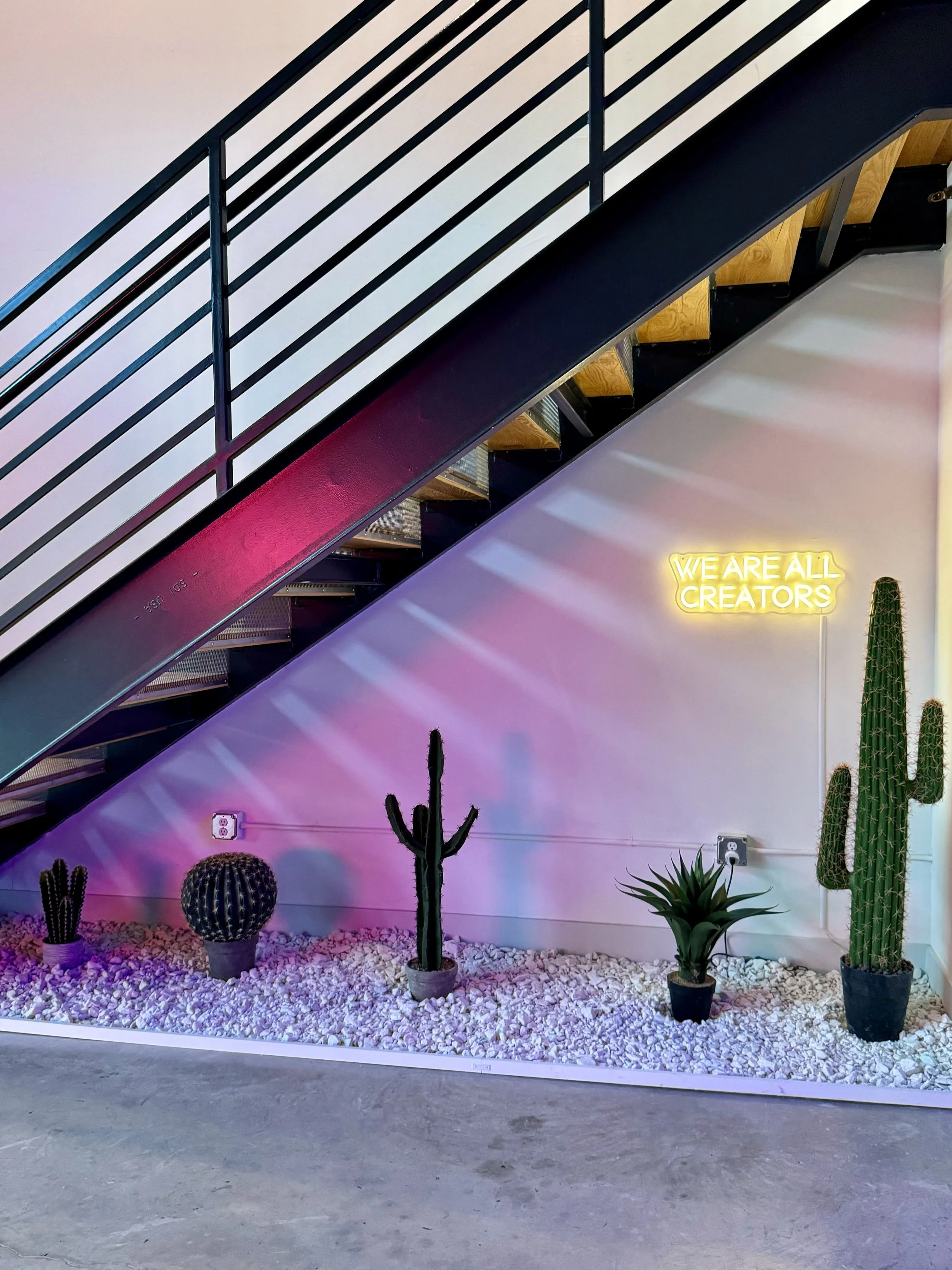 Decorative indoor space with potted cacti and plants beneath a staircase, featuring a neon sign that reads 'We Are All Creators' on a wall illuminated with purple and pink lights.
