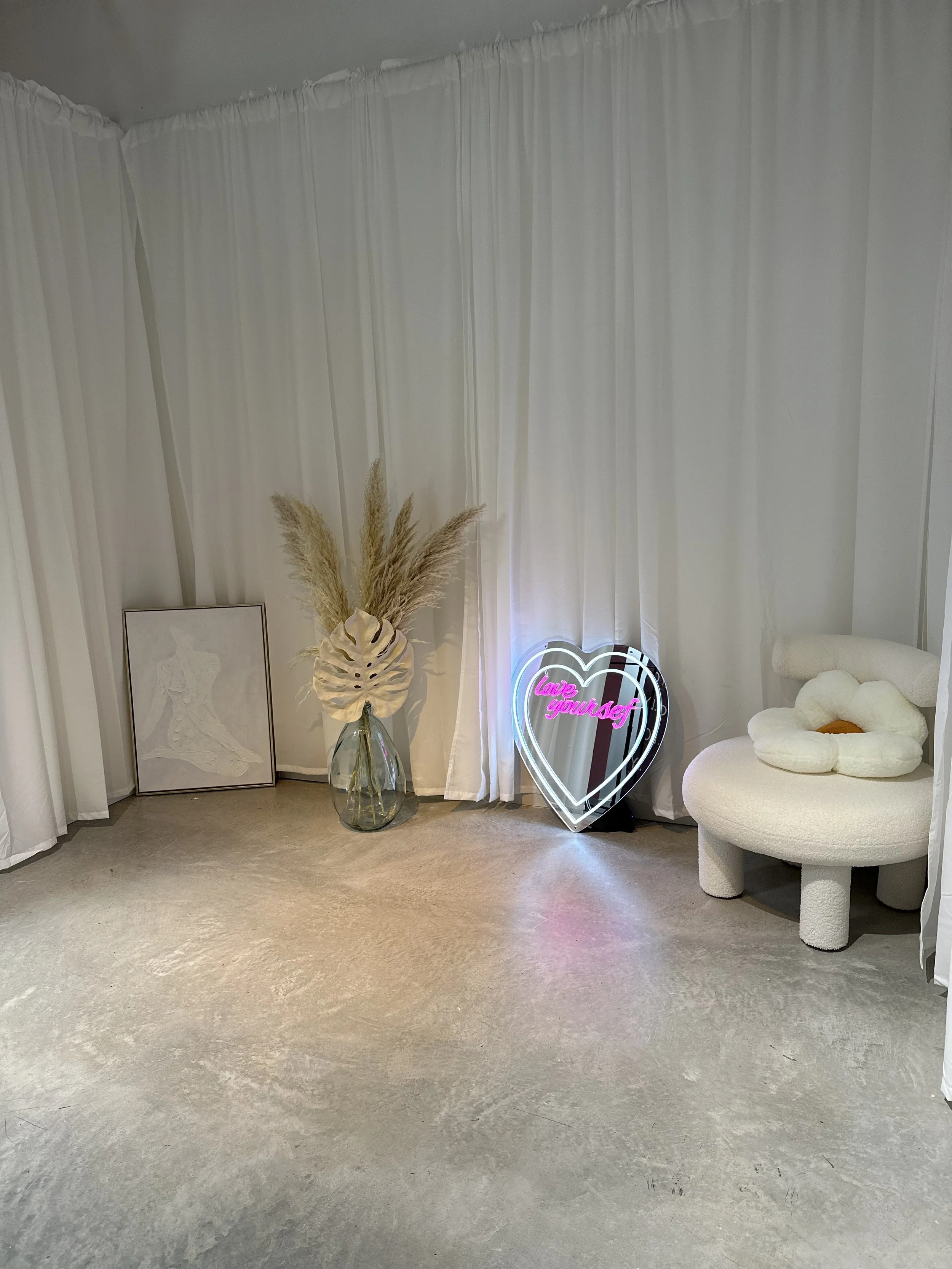 Decorative corner with white curtains, a large glass vase containing pampas grass and a monstera leaf, a framed artwork leaning against the wall, a neon heart-shaped light with the words 'love yourself' in pink, and a modern white armchair with a flo