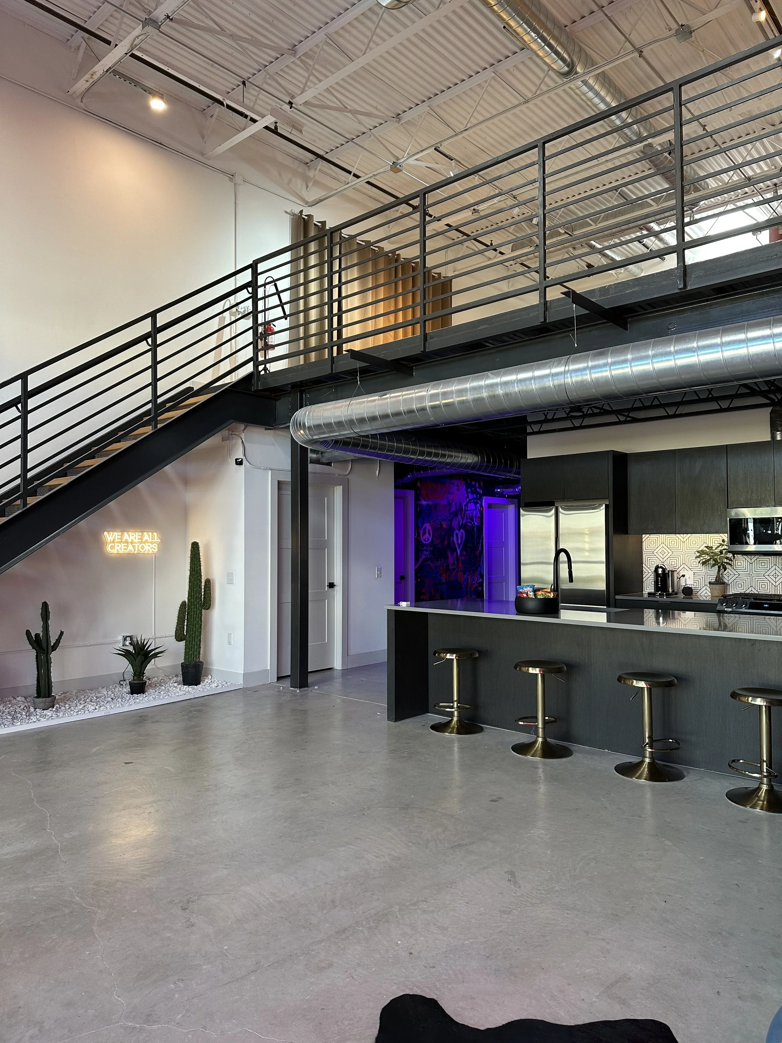 Modern industrial-style open-concept kitchen and living space with concrete floors, black kitchen island with four gold bar stools, stainless steel appliances, and a lofted area with black metal railing and staircase. Decor includes cacti, a neon sig