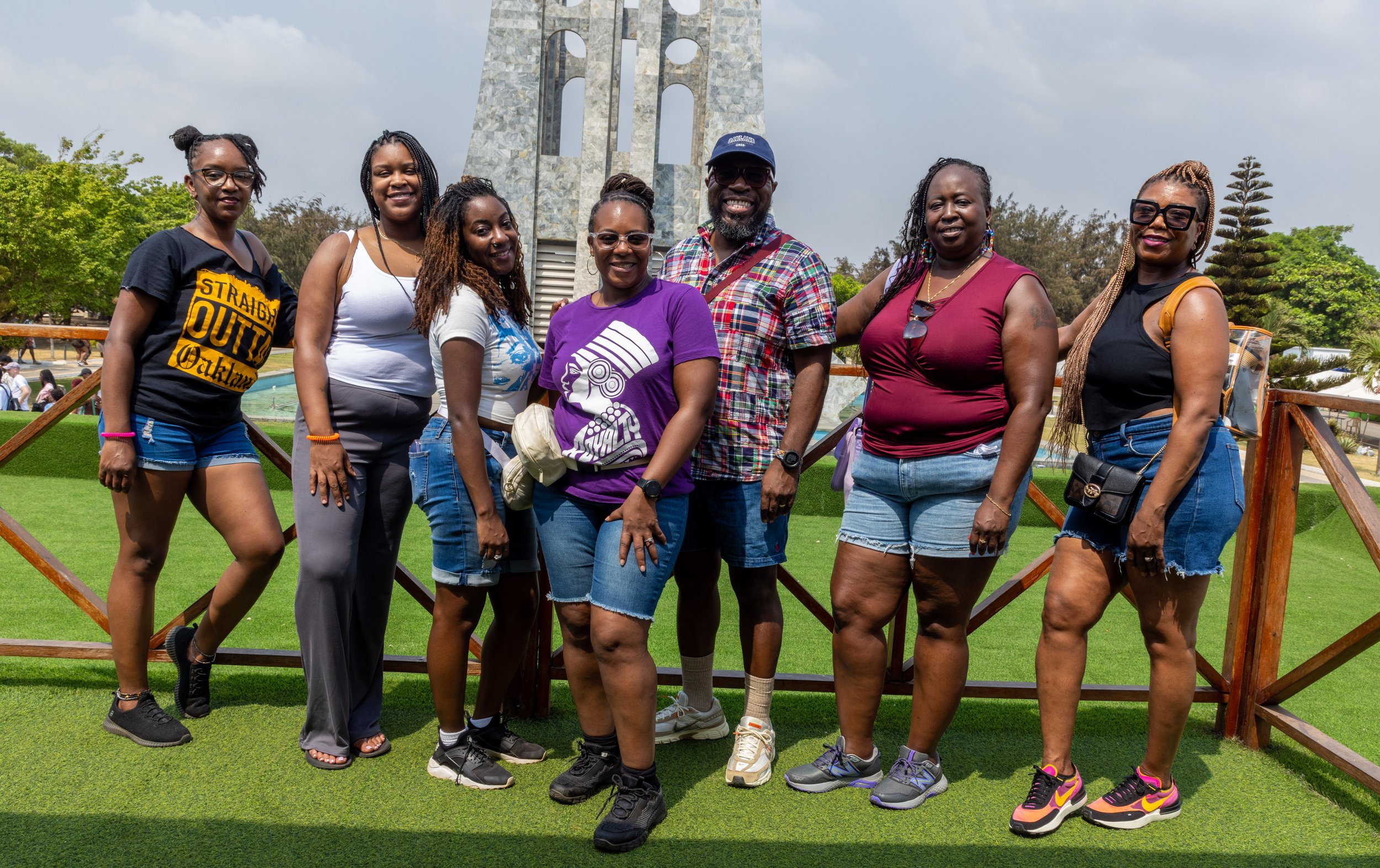 A group of nine diverse people, mostly women, standing outdoors on grass with a monument in the background. They are smiling and dressed casually, some wearing sunglasses and sneakers, with lush green trees around.