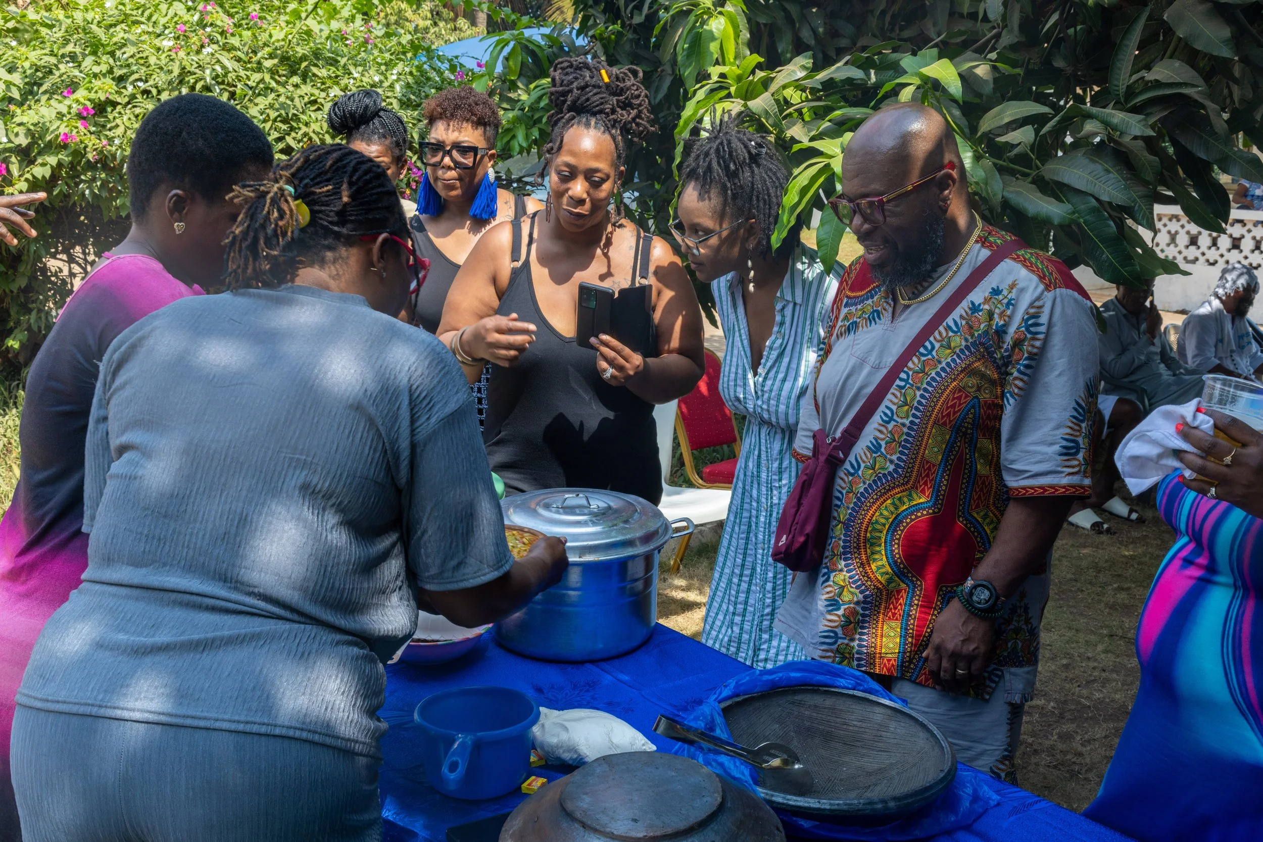 A group of people gathered around a table outdoors, looking at food and a woman serving from a large pot, with others observing and taking photos, surrounded by greenery and some seated in the background.