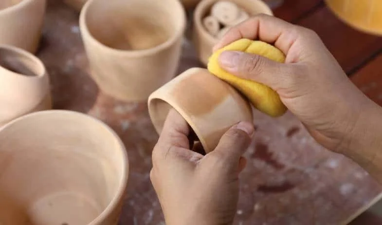 Hands polishing a small clay pot with a yellow sponge, surrounded by other unfinished clay pots on a work table.