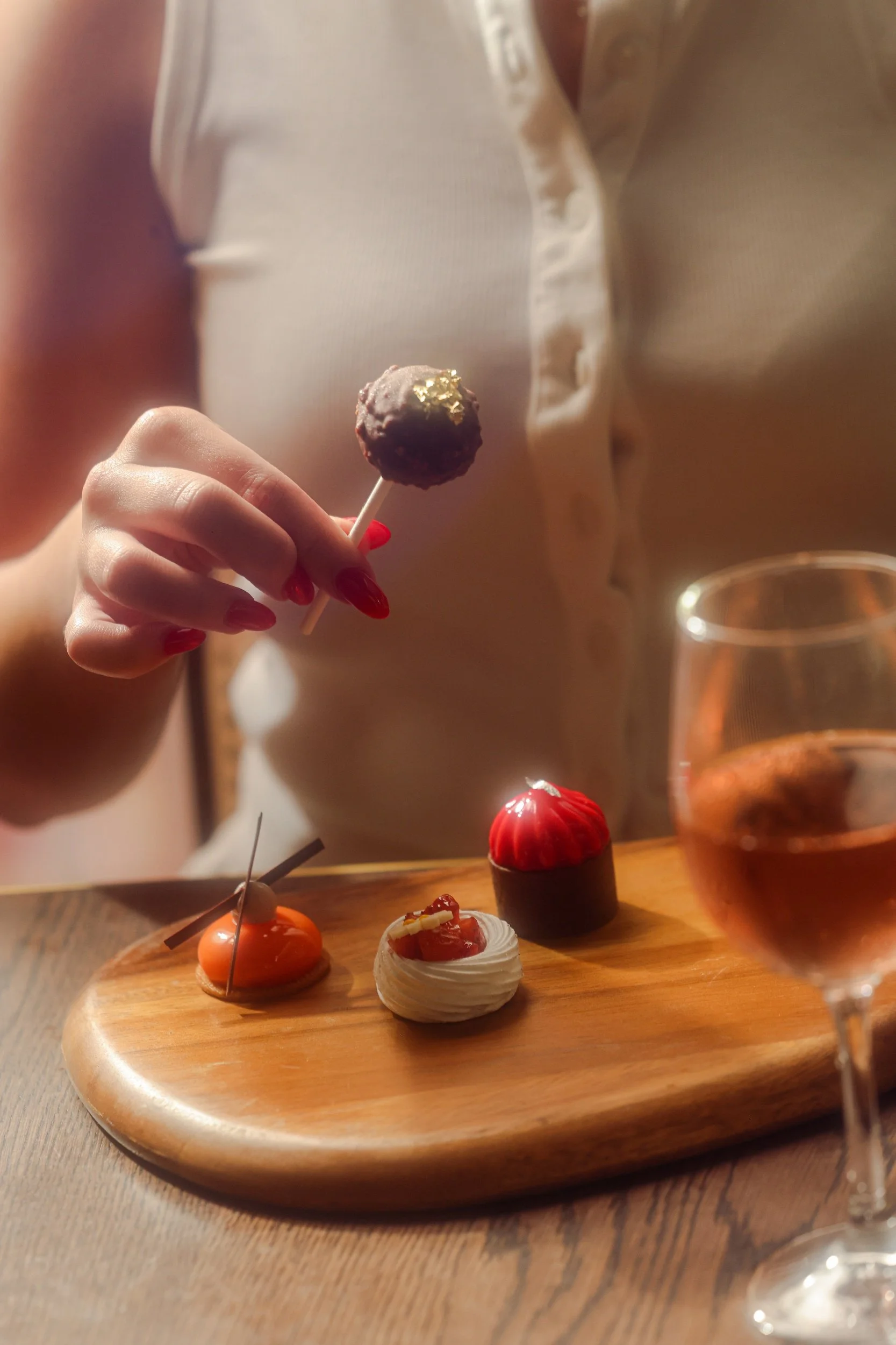 Person with red painted nails holding a chocolate cake pop with gold leaf, surrounded by small decorative desserts on a wooden serving board, and a glass of rose wine in the foreground.