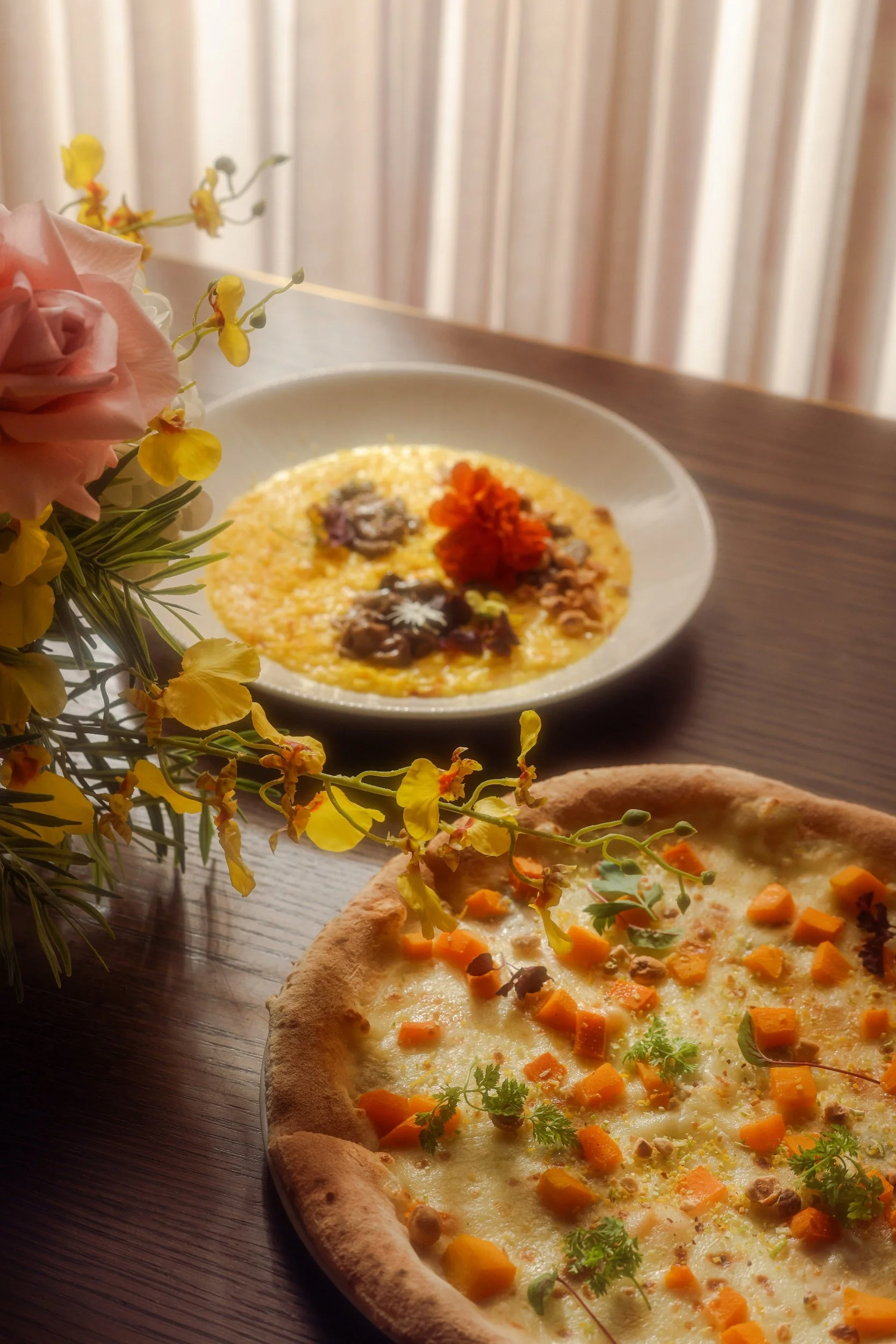 A pizza with cheese, diced butternut squash, and herbs on a wooden table. In the background, a bowl of rice topped with flowers and garnishes, and a bouquet of flowers on the side.