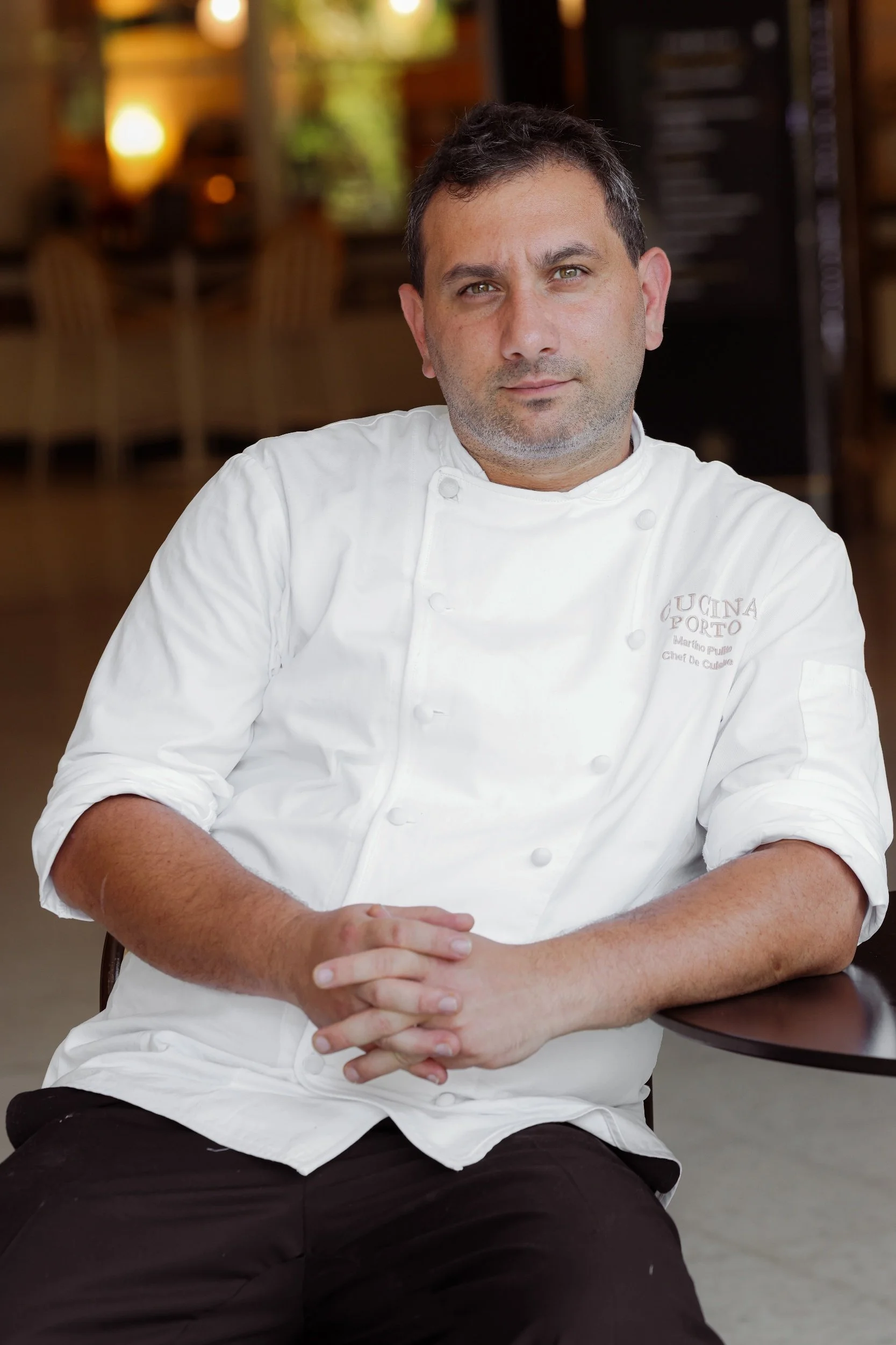 A male chef wearing a white chef's coat sits at a table, with hands clasped, in a restaurant setting.