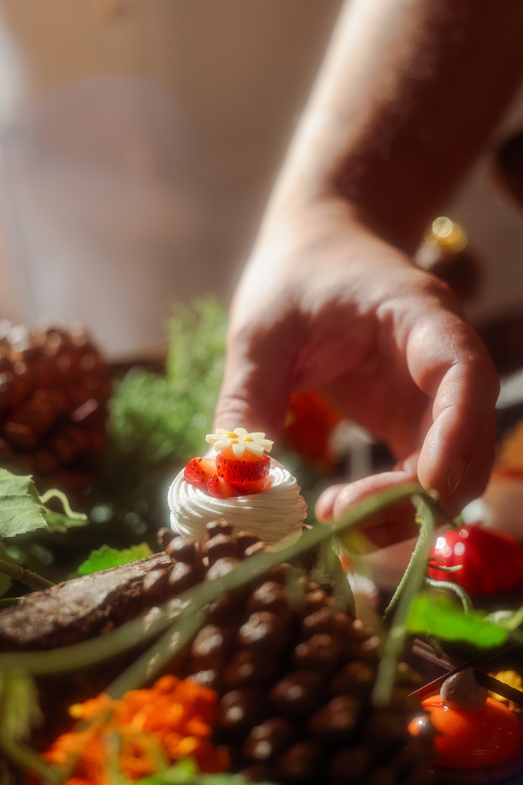 Close-up of a person arranging a small dessert topped with strawberries and a daisy-shaped edible decoration, surrounded by pinecones, green leaves, and other colorful elements.
