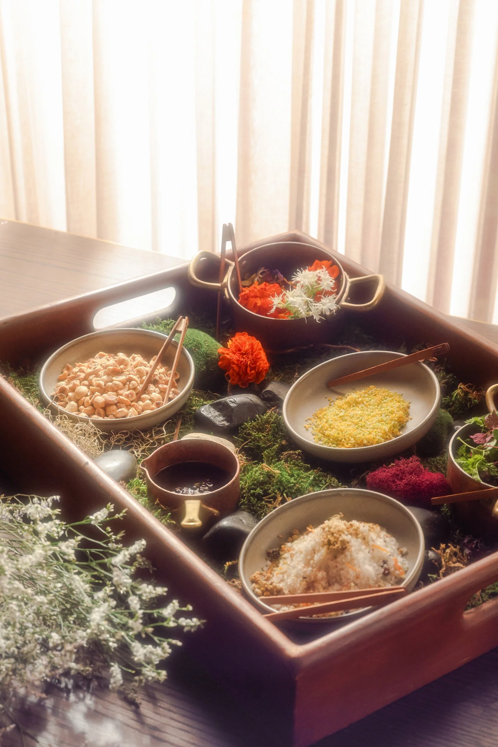 A wooden tray holding various small bowls with different ingredients or spices, decorated with flowers and moss, placed on a wooden table with light-colored curtains in the background.