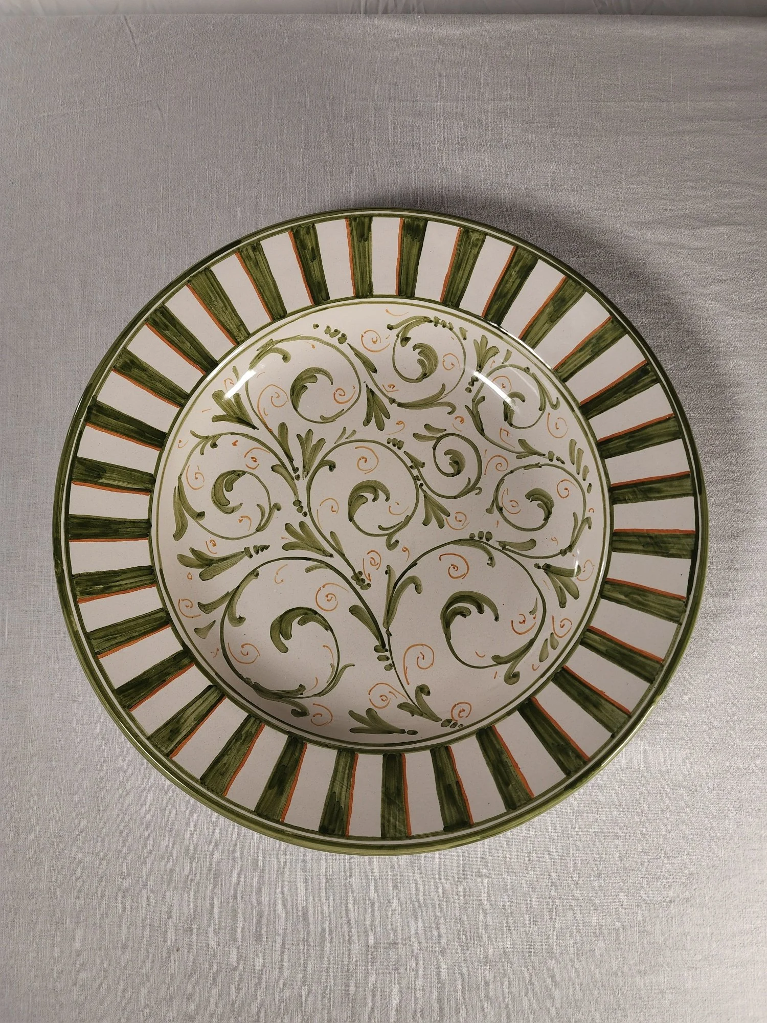 Decorative ceramic plate with a white background, green swirling floral pattern, and a border featuring green and orange stripes, on a light-colored fabric surface.