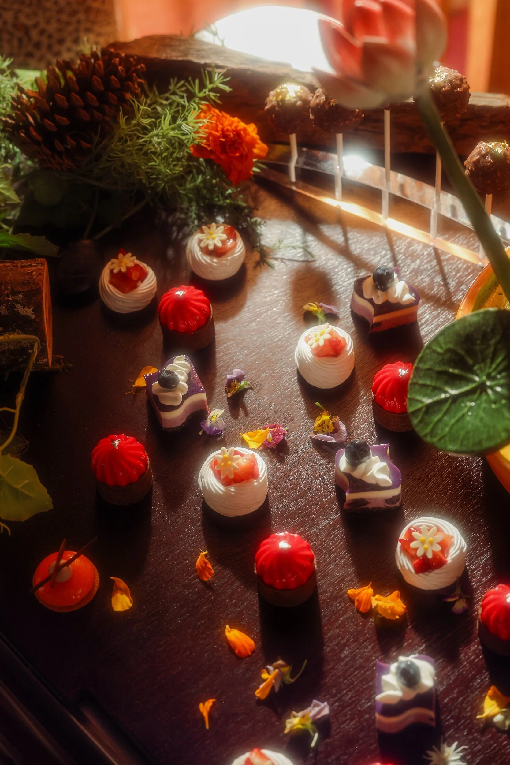 Assorted miniature desserts on a wooden surface, decorated with edible flowers, surrounded by greenery and a log, with warm sunlight.
