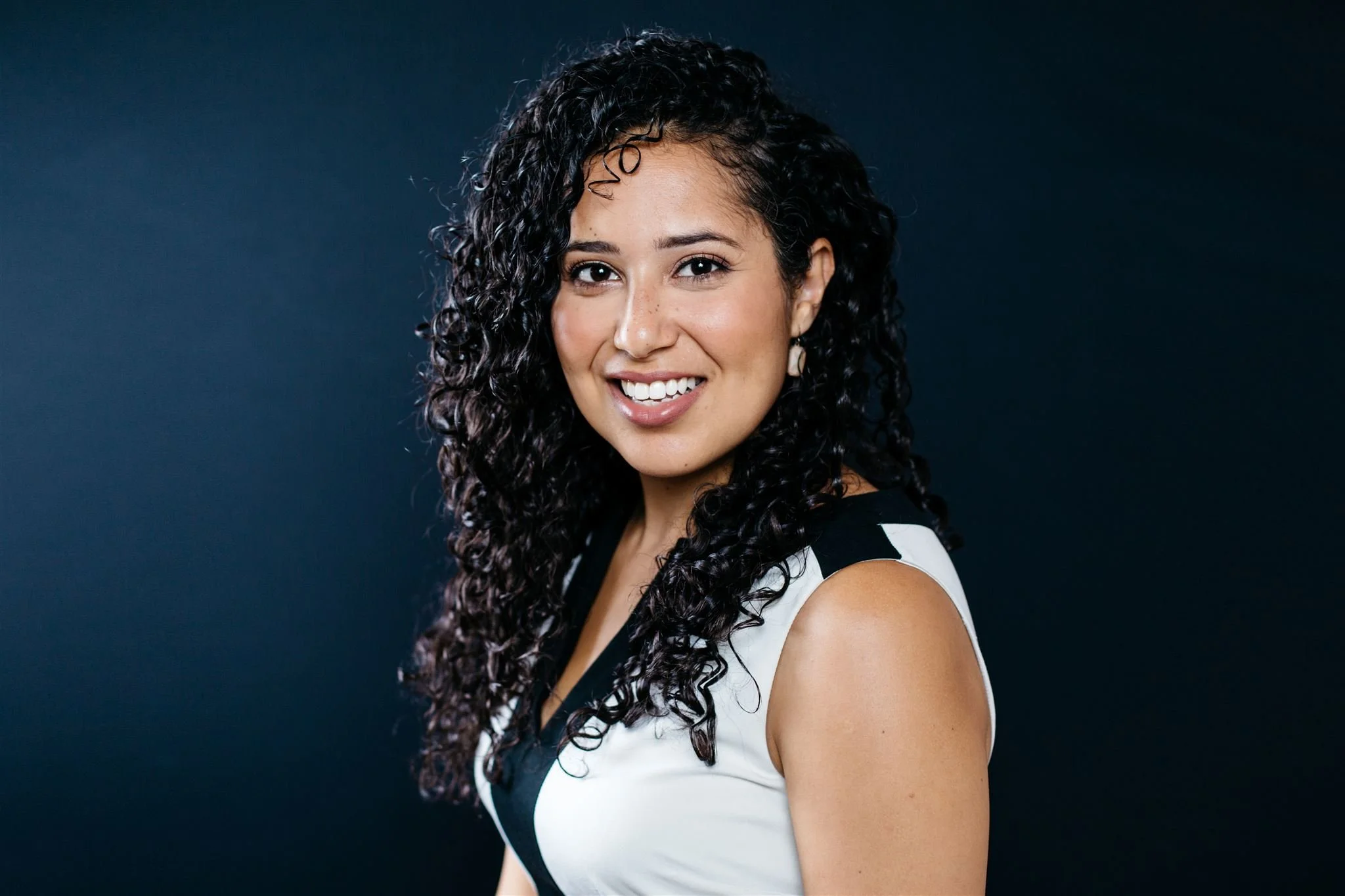 Actor headshot of a smiling woman with long dark curls in a black-and-white sleeveless top, Montreal