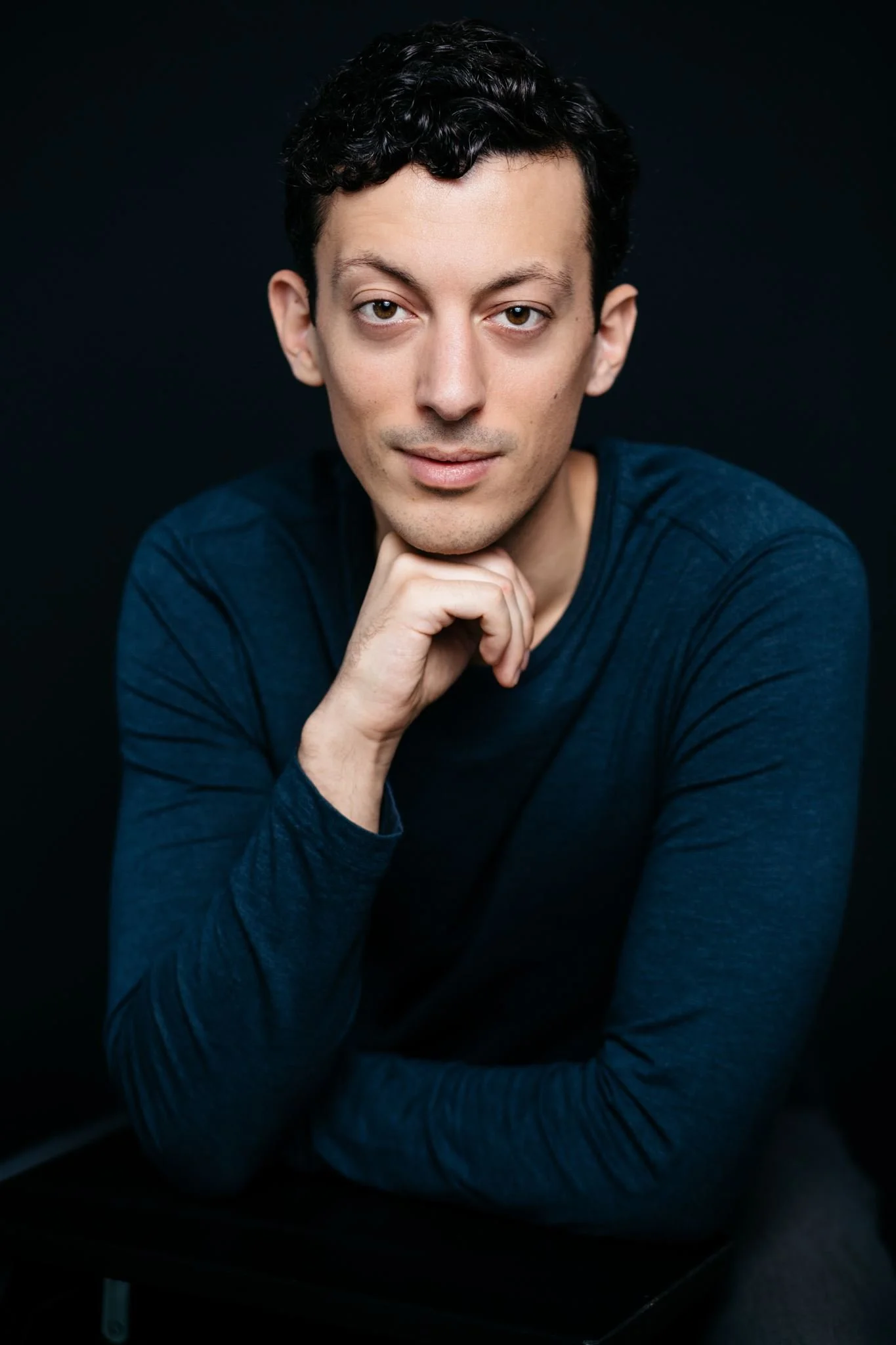 Man with short dark curly hair, teal sweater, hand at chin, dark background - actor headshot by Tea & Oranges, Montreal portrait photographer
