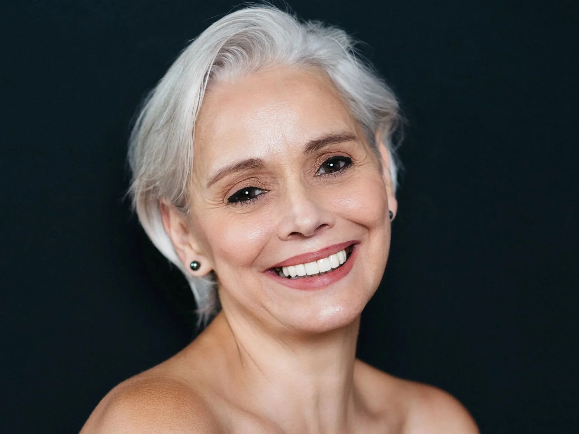 Actor headshot of a beaming woman with short silver hair and bare shoulders on a black backdrop, Montreal