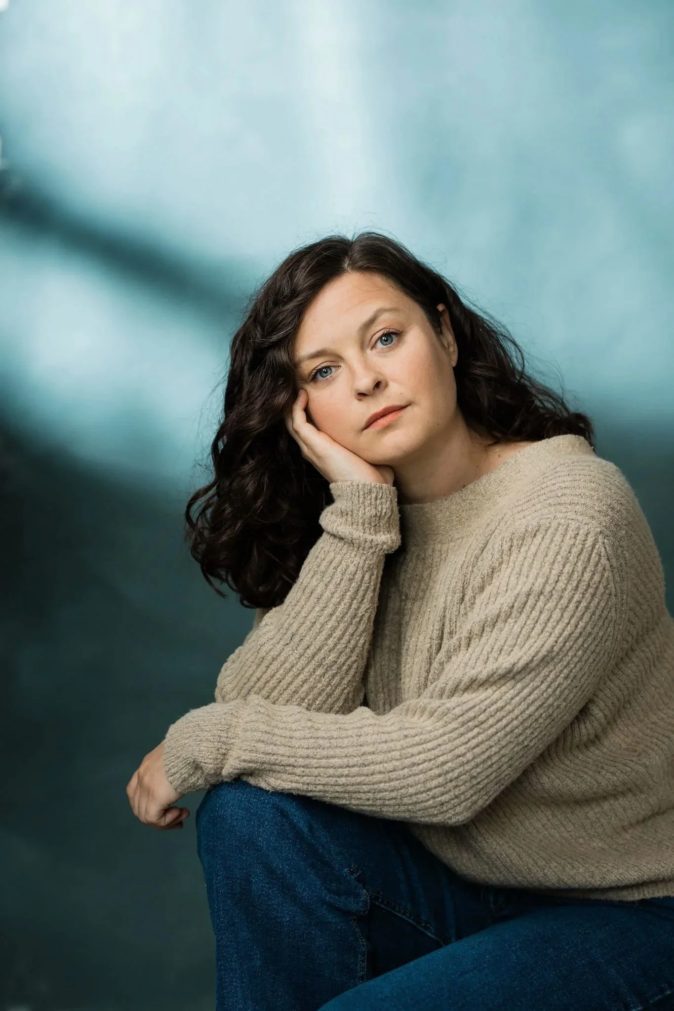 Photo de casting d'une comédienne aux boucles brunes en pull beige, joue appuyée sur la main, fond bleu-vert, Montréal