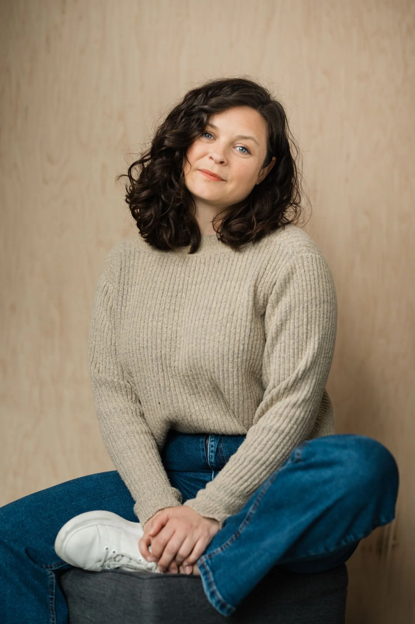 Photo de casting d'une comédienne aux cheveux bruns ondulés, pull beige et jean, assise jambes croisées, Montréal