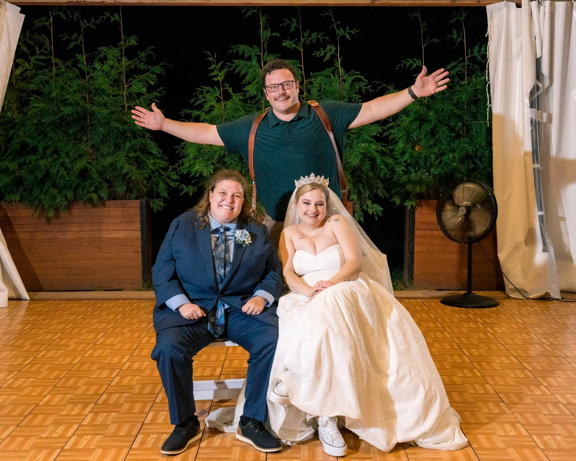 Three people at a wedding event, with a woman in a wedding dress and tiara sitting in the middle, a person in a suit sitting beside her, and a man standing behind with arms outstretched.