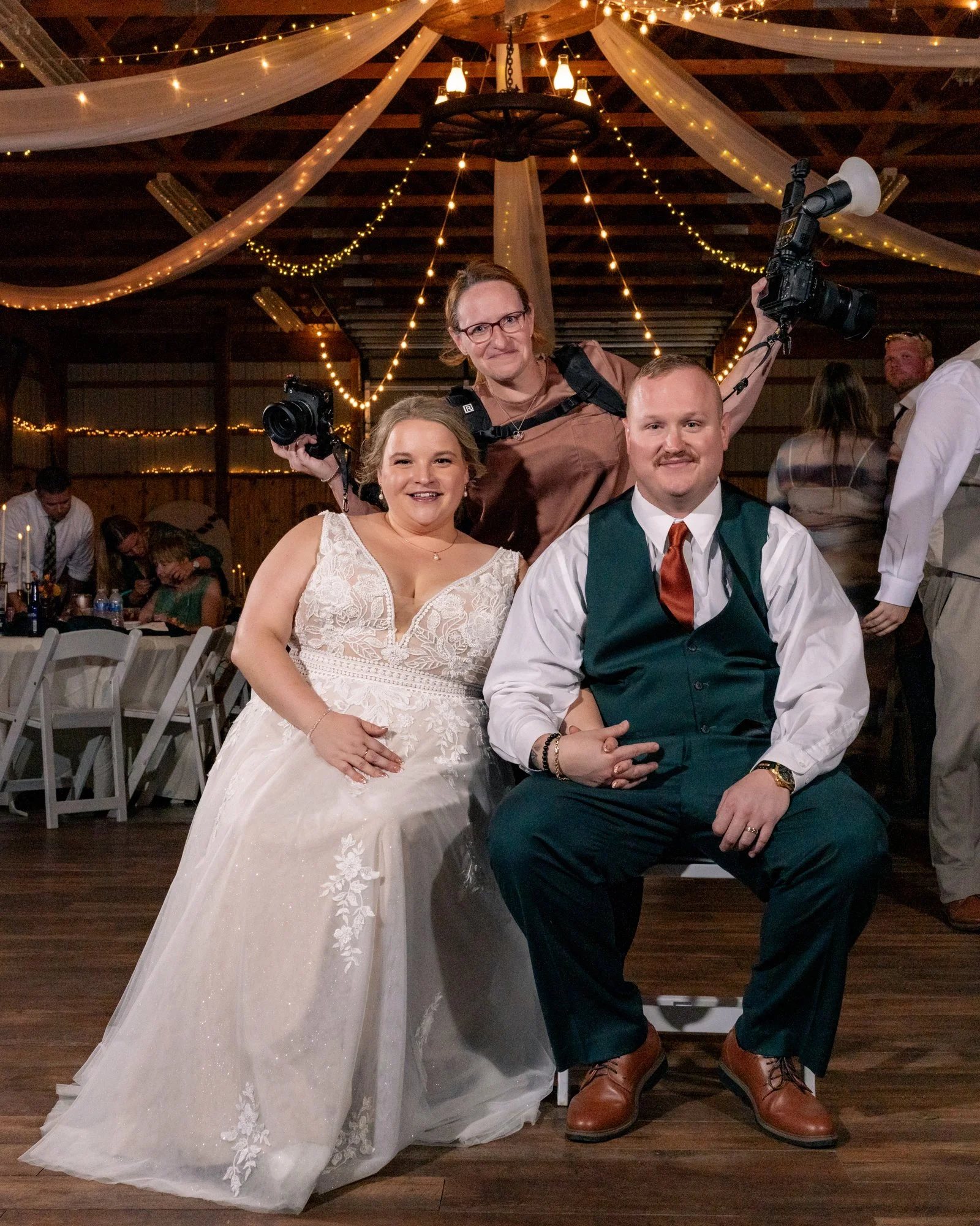 A bride and groom at a wedding reception, seated while a woman stands behind them. The bride is in a white lace wedding dress, and the groom is in a dark green vest and tie. The woman behind them holds cameras and has cameras around her neck. The reception room is decorated with string lights and draped fabric, with people sitting at tables in the background.
