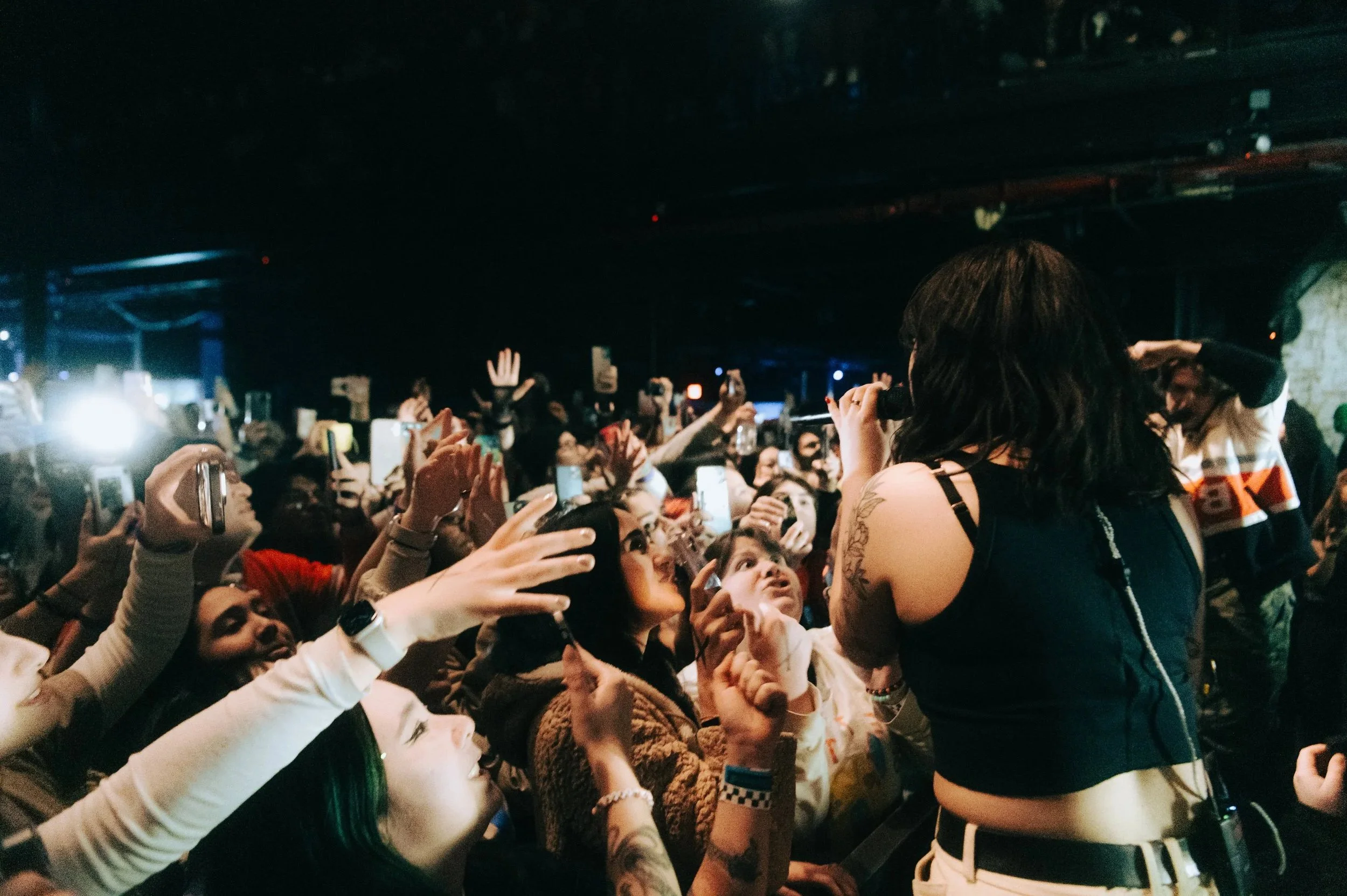 Fans crowd around mxmtoon at a Ricky Montgomery concert, many holding up phones to take pictures or videos, with a performer on stage in the foreground.