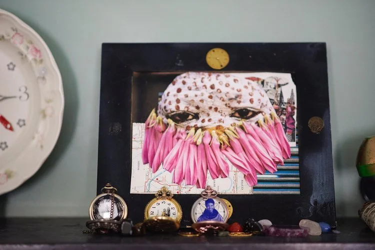 Decorative collage art of a face with large eyes, surrounded by pink petal-like shapes, framed on a black background, with pocket watches and small objects on a shelf and a clock on the wall to the left.