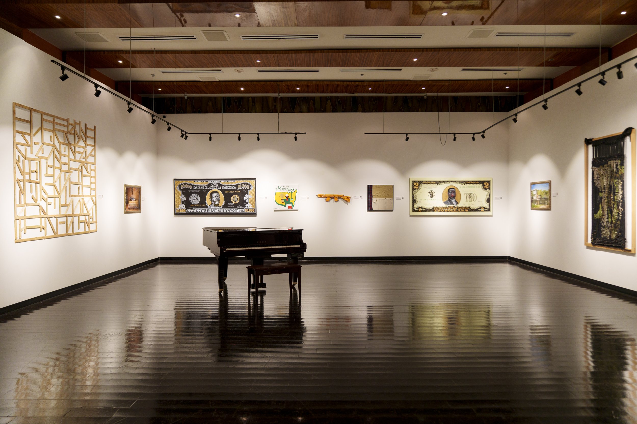 An art gallery with white walls and wooden ceiling, displaying various artworks including paintings, mixed media pieces, and sculptures. A black piano and bench are at the center of the room, reflecting on the dark wooden floor. Track lighting illumi