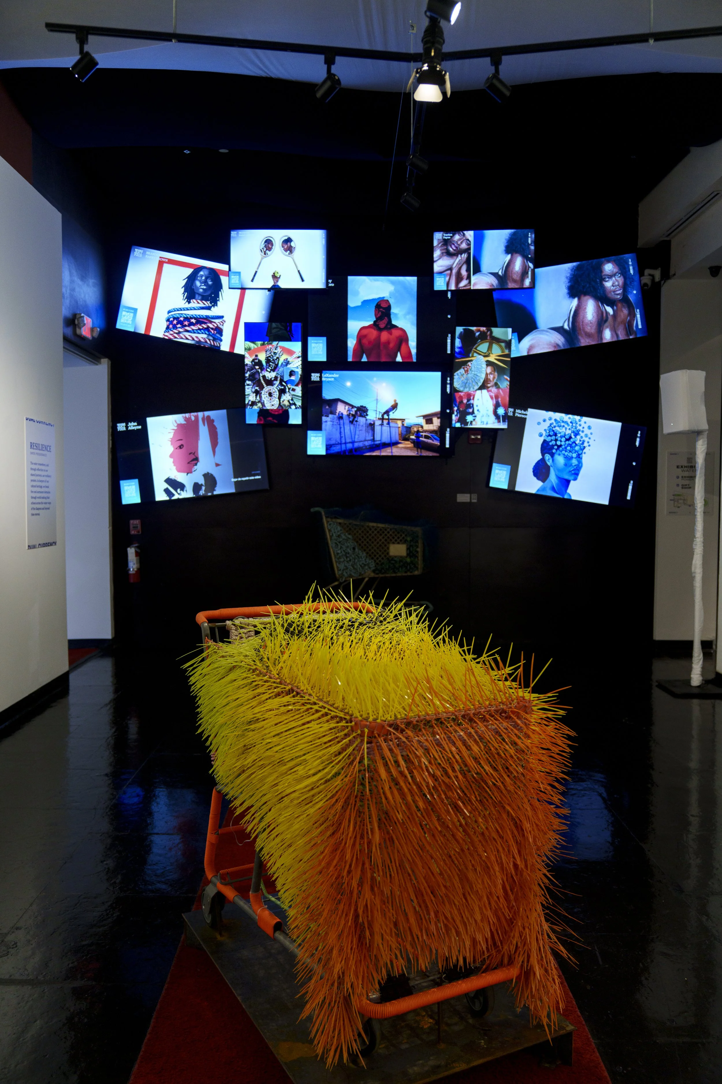 An art installation featuring a shopping cart covered in bright yellow and orange spikes, positioned in front of a wall with multiple digital screens displaying colorful artwork and photographs.