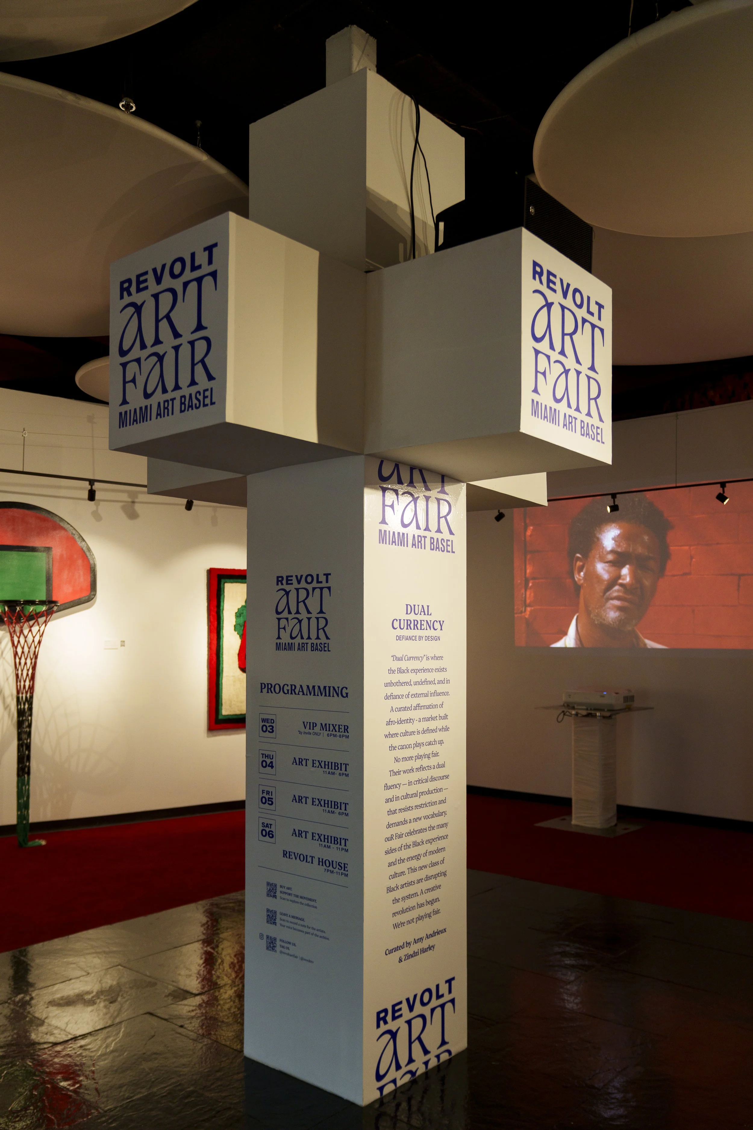 Exhibit pillar at Revolt Art Fair in Miami with event schedule and description, with a basketball hoop and artwork visible in the background.