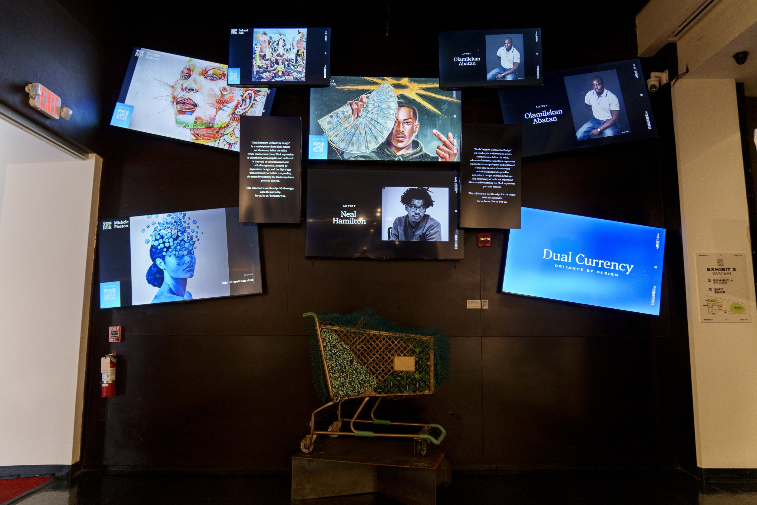 Multi-screen display wall featuring various digital artworks, artist portraits, and informational text in a gallery setting, with a shopping cart in front.
