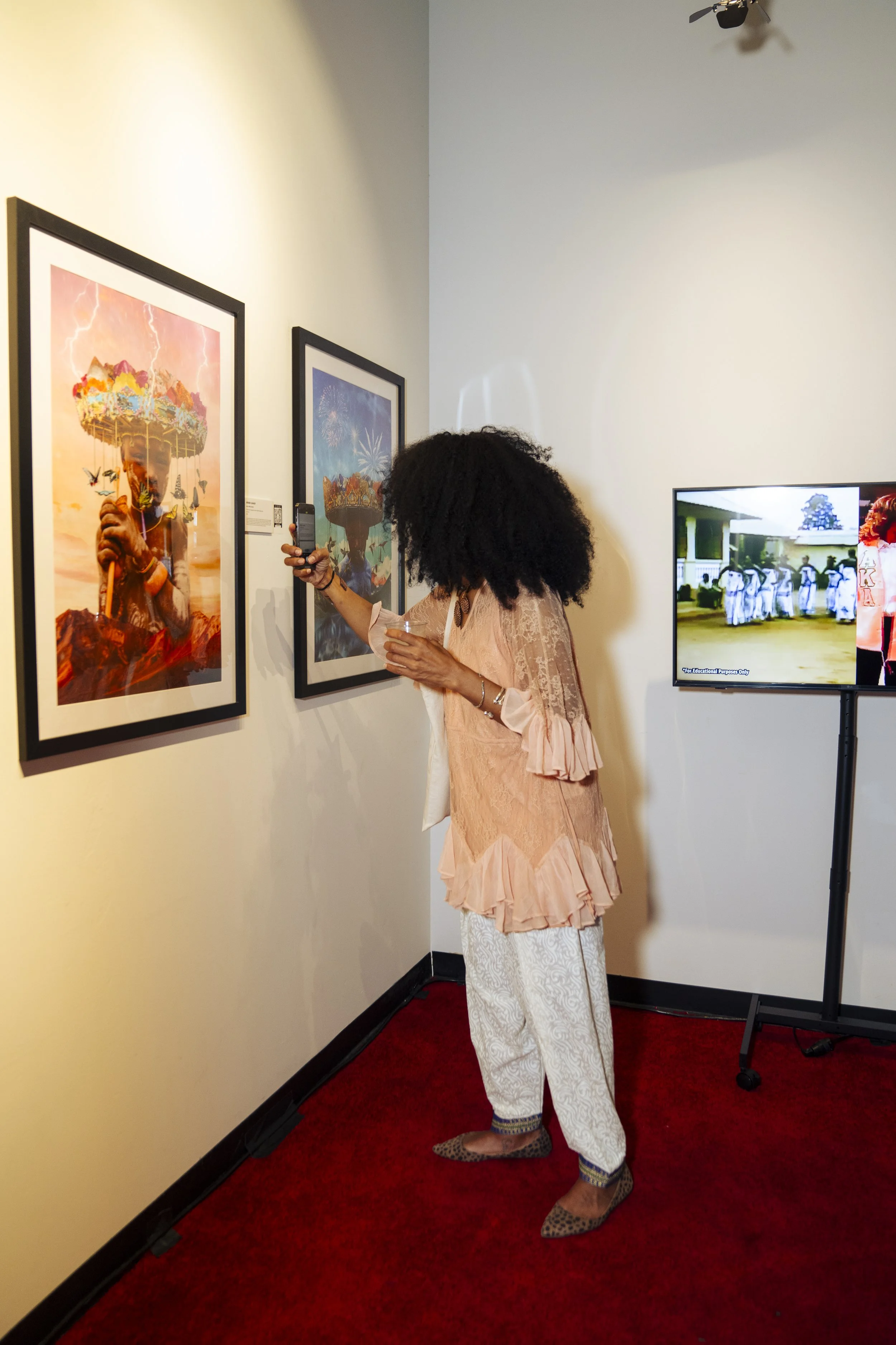 A woman with curly hair, wearing a pink lace blouse and white patterned pants, is taking a photo of framed artwork at an art gallery.