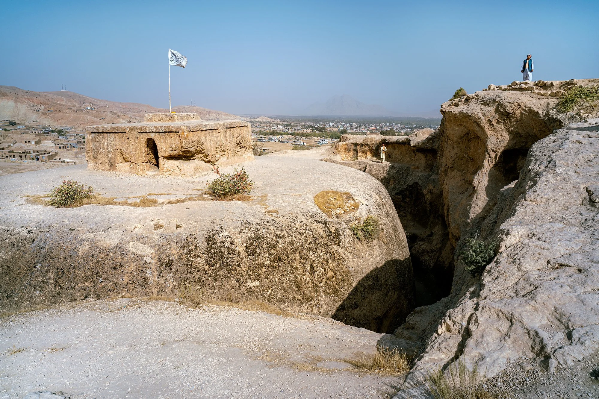 The Taliban flag flies over the historic site of Takht-e Rostam, a Buddhist stupa carved into the rock that has survived centuries of history. The contemporary power proclaims itself the guardian of a past that precedes it, imposing its authority ove