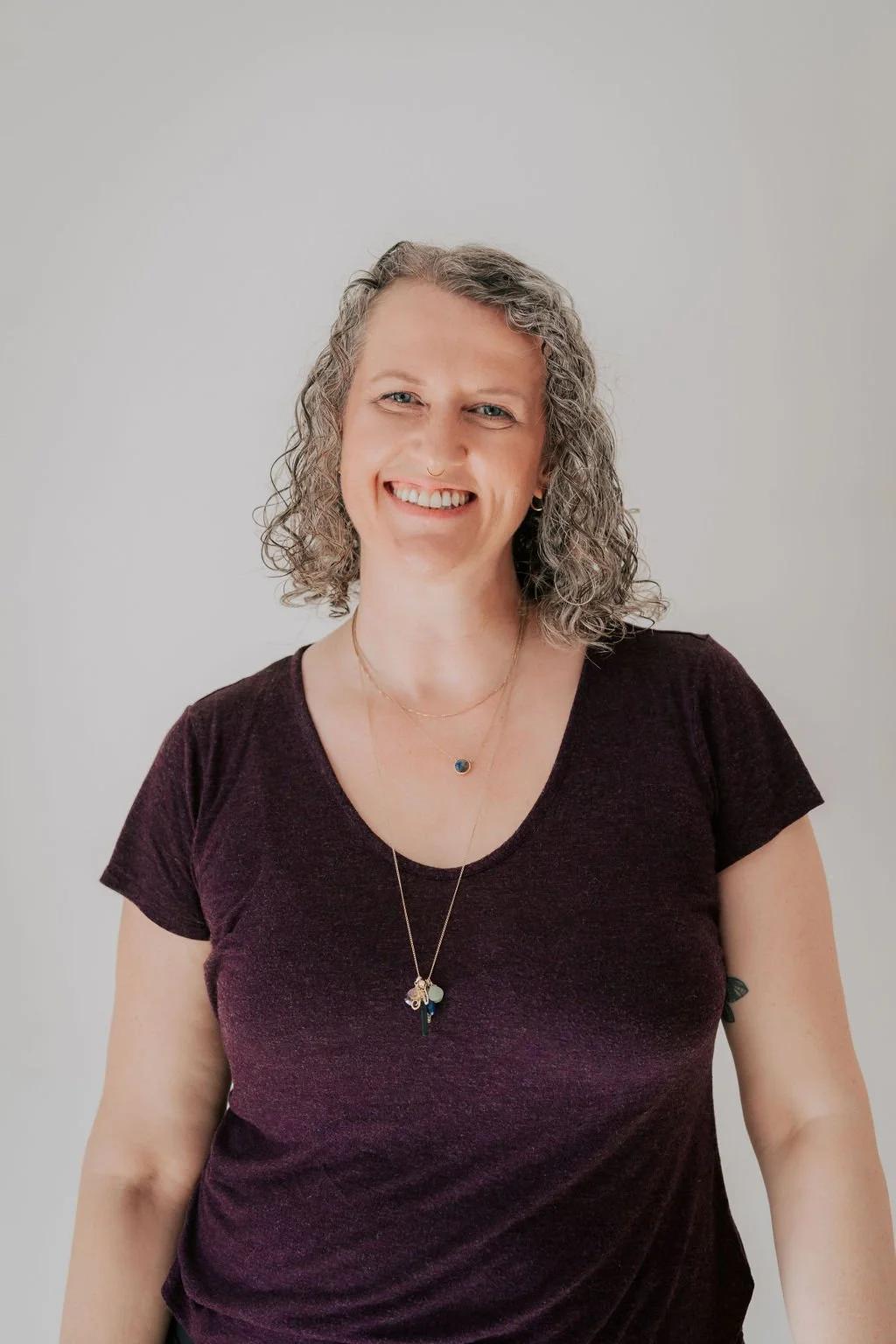 A smiling woman with curly gray hair wearing a dark purple t-shirt and layered necklaces standing against a plain light background.