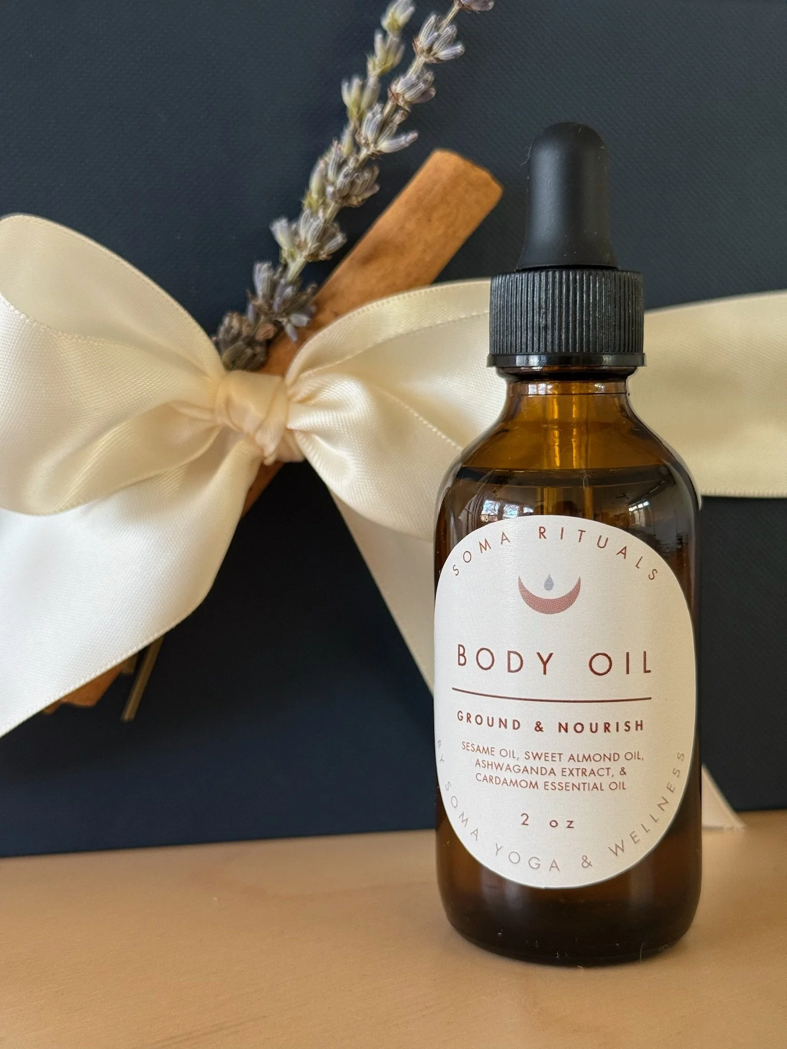 Amber glass bottle of Soma Rituals Body Oil with dropper cap, label reads 'Ground & Nourish' with ingredients listed, positioned on a wooden surface beside a gift wrapped with a cream satin ribbon, lavender sprig, and cinnamon stick.