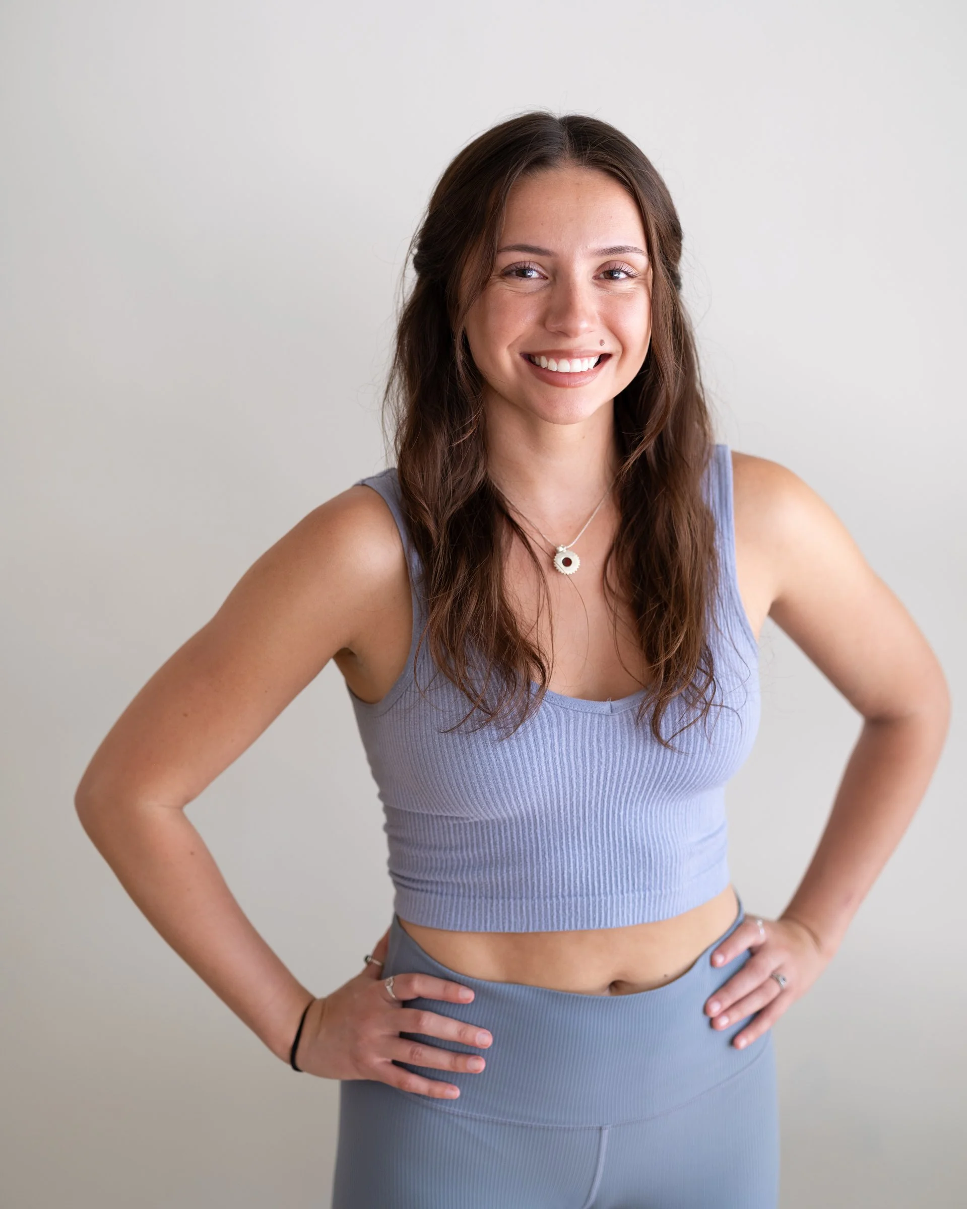 A young woman with long, wavy brown hair wearing a light purple sleeveless crop top and matching high-waisted leggings, smiling with her hands on her hips against a plain gray background.